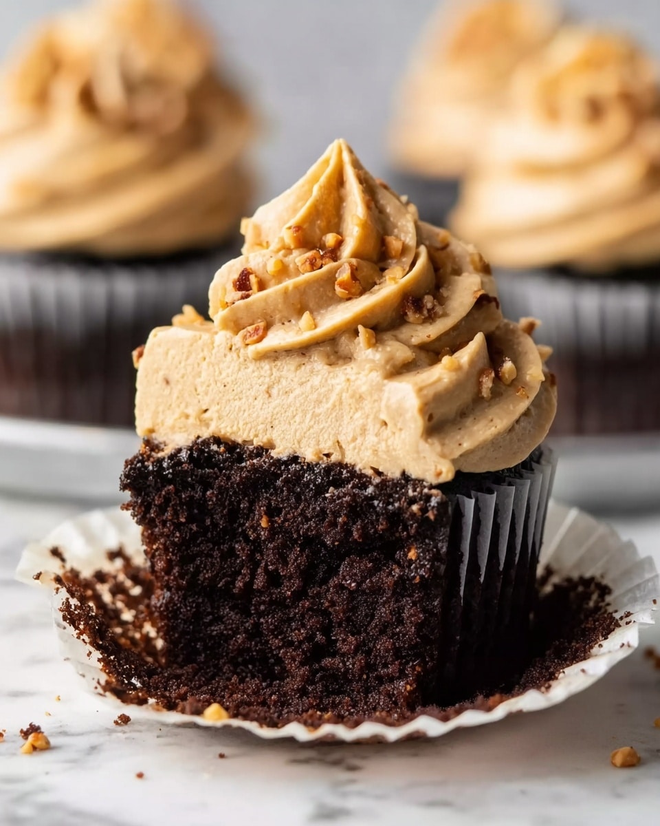 A close-up view of a cupcake with two clear layers, sitting on a white marbled surface. The bottom layer is a dark brown, moist chocolate cake with a soft, crumbly texture. The top layer is thick and creamy light brown frosting swirled into a tall peak with small nut pieces sprinkled inside. The cupcake wrapper is white with some chocolate crumbs scattered around it. In the background, there are blurred cupcakes with the same frosting, all set on the same white marbled surface. Photo taken with an iphone --ar 4:5 --v 7