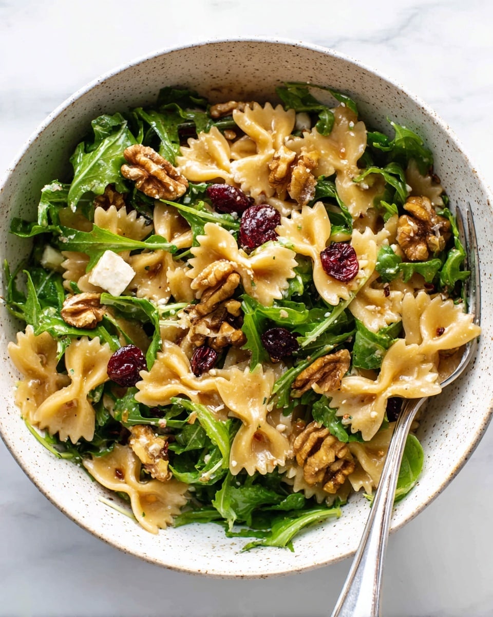 A white speckled bowl filled with a pasta salad showing one layer of farfalle pasta in light golden color, mixed with fresh green arugula leaves scattered all over. The salad includes pieces of light beige cheese cubes and brown walnut halves placed on top and within the mix. Small dark red dried cranberries are spread throughout, adding color contrast to the dish. A silver fork is partially inserted into the salad on the right side of the bowl, resting on the food. This is all set on a white marbled surface. Photo taken with an iphone --ar 4:5 --v 7