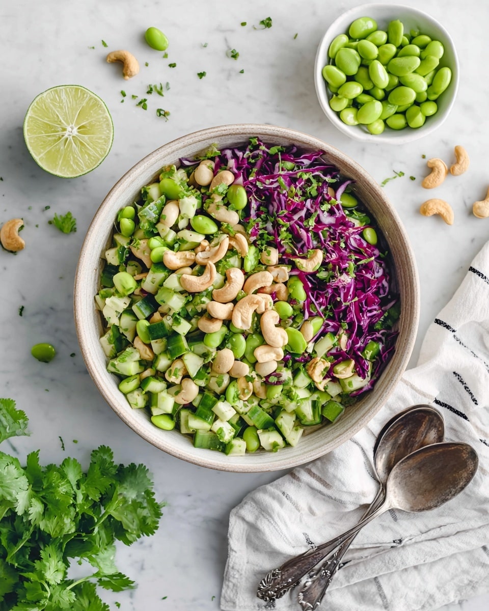 A bowl of fresh salad sits at the center on a white marbled surface, filled with three main layers: the bottom layer is light green diced cucumber pieces, the middle layer has bright green edamame beans and white beans scattered throughout, and the top layer includes thin strips of purple cabbage and small chopped green herbs sprinkled evenly. Cashew nuts are spread on top, adding a light beige color and chunky texture. On the white marbled surface around the bowl, there is a half lime, green cilantro leaves, and some extra edamame beans in a white bowl. Two silver spoons rest on a white cloth with thin black stripes near the bottom right. The scene is bright with natural light and a clean, fresh look. photo taken with an iphone --ar 4:5 --v 7