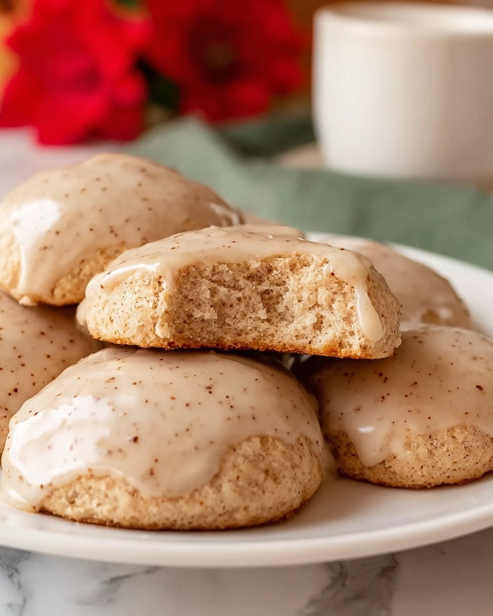 The image shows a close-up of several soft, light brown biscuits on a white plate. Each biscuit has a smooth, slightly shiny glaze with small specks of spice visible on the surface. One biscuit is stacked slightly on top of others and has a bite taken from it, revealing a light, fluffy inside. The plate sits on a white marbled surface. In the blurred background, there is a red flower and part of a white cup. The overall look is warm and inviting, with a cozy feel. photo taken with an iphone --ar 4:5 --v 7
