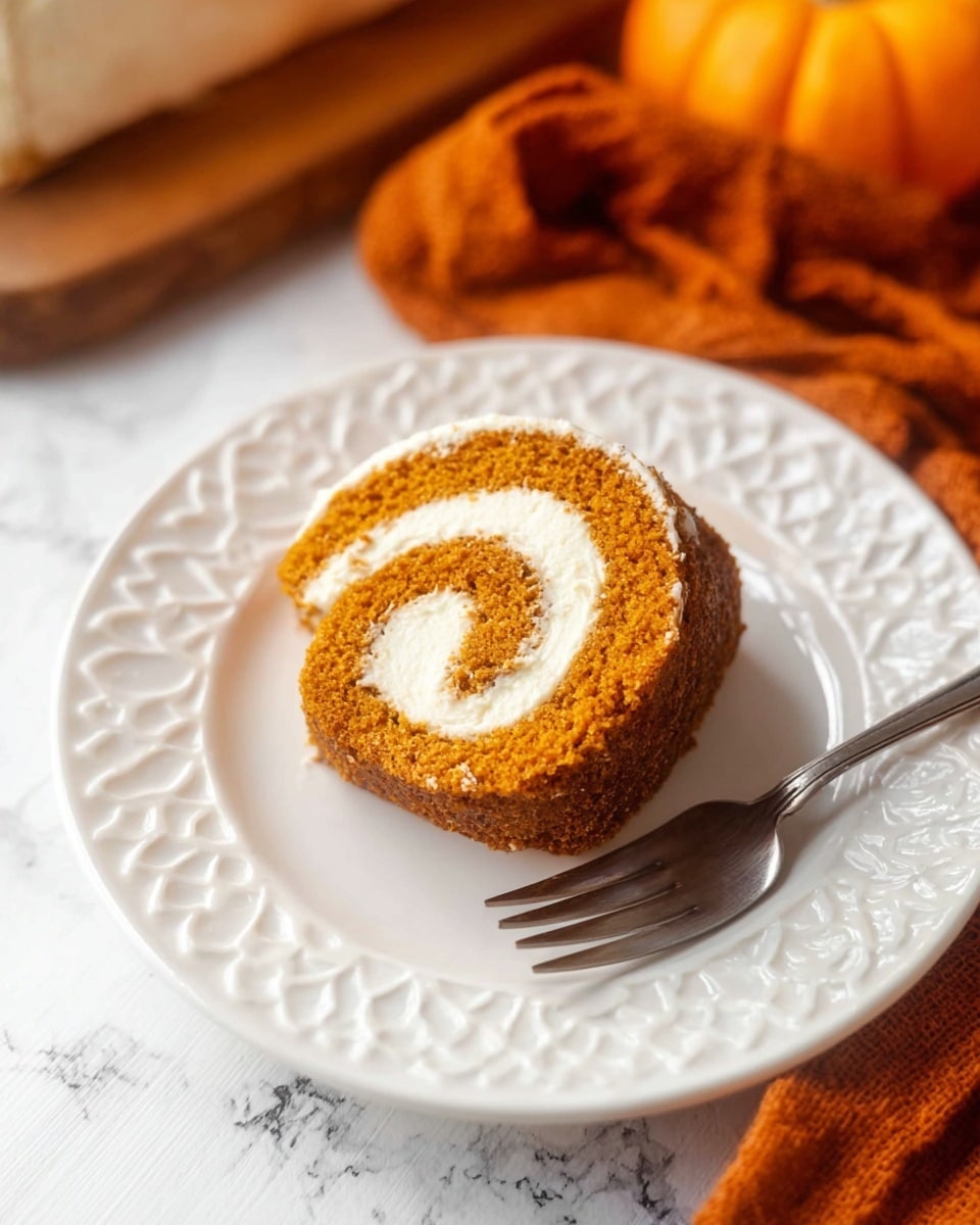 A single slice of a pumpkin roll cake sits in the center of a white plate with a raised, ornate pattern along the edge. The cake has two layers: an outer soft and spongy orange layer, and an inner smooth pale cream filling, rolled in a spiral shape. A silver fork rests to the right side of the slice on the plate. In the background, a folded burnt orange fabric is partially visible, along with a hint of a pumpkin, all against a white marbled surface photo taken with an iphone --ar 4:5 --v 7
