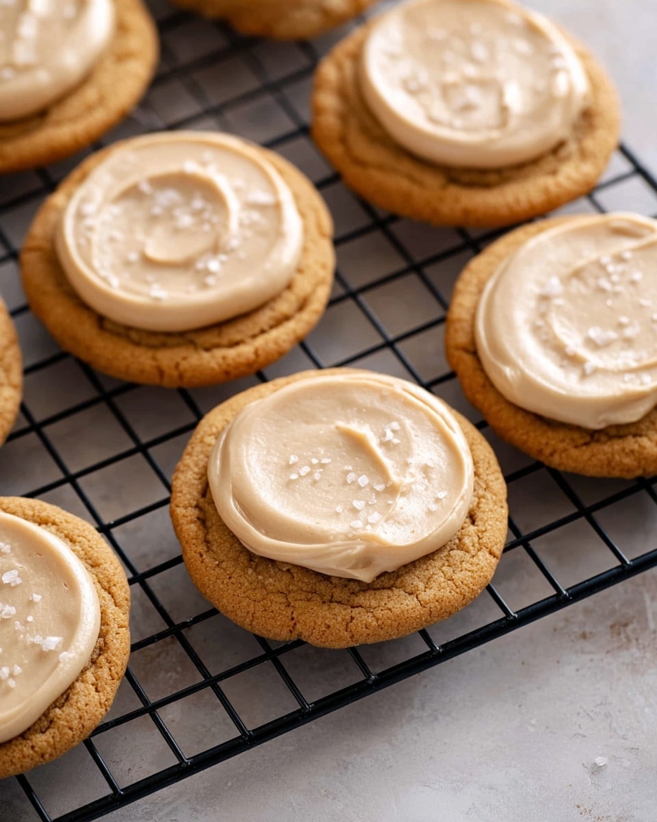 The image shows six round cookies placed on a black wire cooling rack, each topped with one layer of smooth, creamy light beige frosting spread in a circular motion. The cookies themselves are golden brown with a slightly cracked surface texture, and the frosting appears soft and glossy. Some white coarse salt flakes are lightly sprinkled on top of the frosting, adding subtle texture contrast. The cookies rest on a white marbled textured surface. The photo taken with an iphone --ar 4:5 --v 7