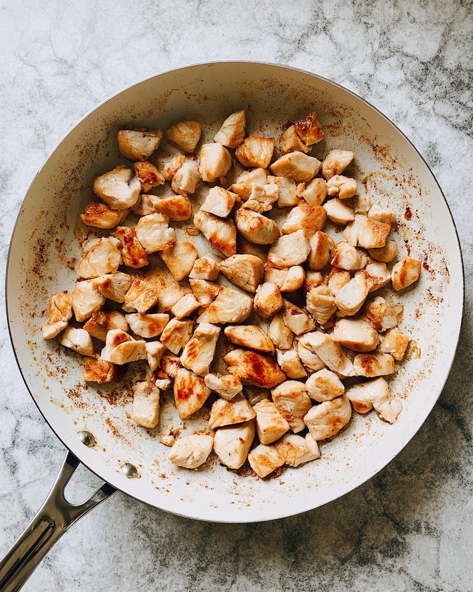 A round white frying pan filled with many small to medium pieces of cooked chicken, lightly browned with a mix of golden and white colors on the pieces. The pan surface shows light browning and some residue from cooking, with a metal handle extending to the right. The pan rests on a white marbled textured surface. photo taken with an iphone --ar 4:5 --v 7