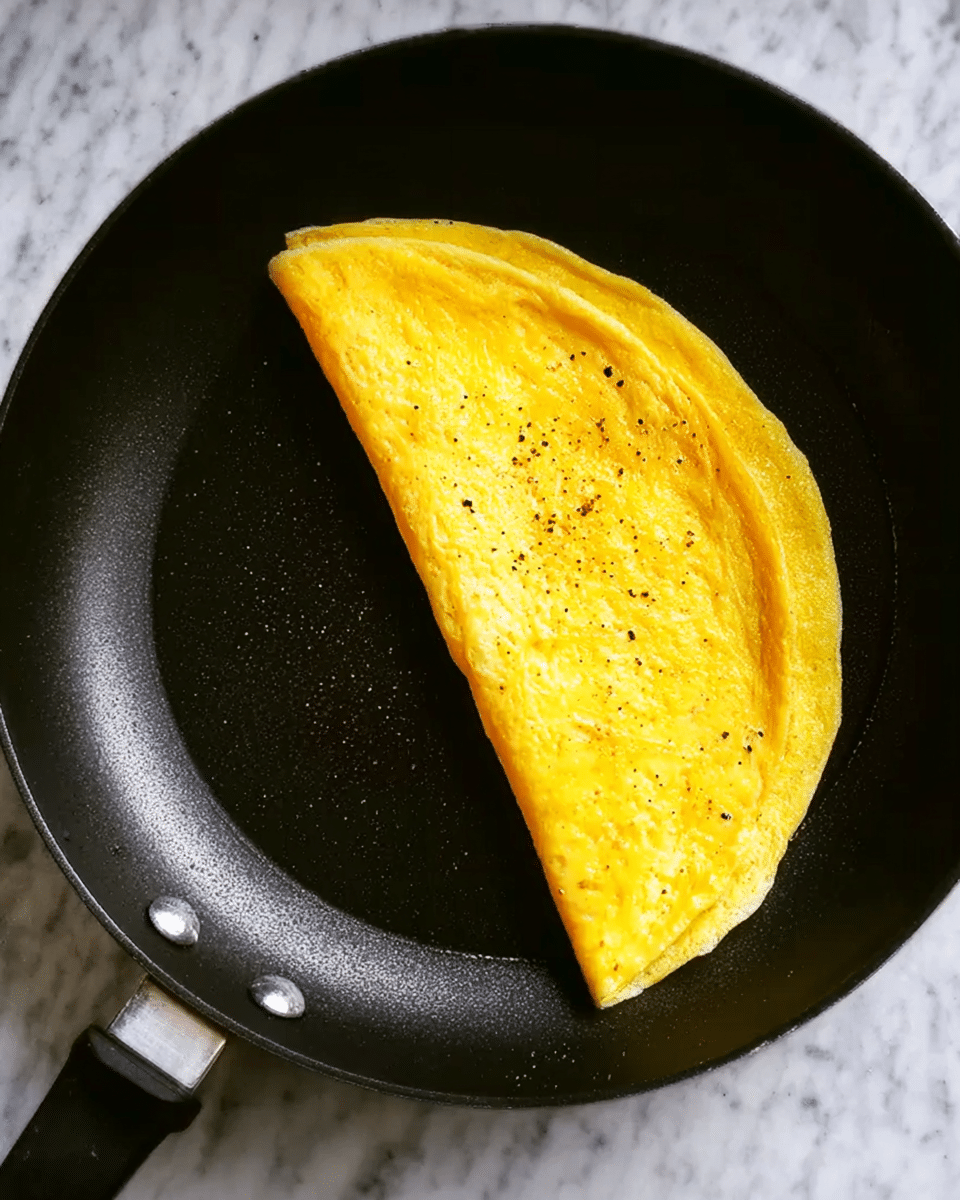 A bright yellow omelette folded in half with a smooth and soft texture, sitting neatly on one side of a black frying pan. The omelette has small black pepper specks on its surface, adding slight contrast. The frying pan has a shiny, slightly oily look, and is placed on a white marbled textured background. Photo taken with an iphone --ar 4:5 --v 7