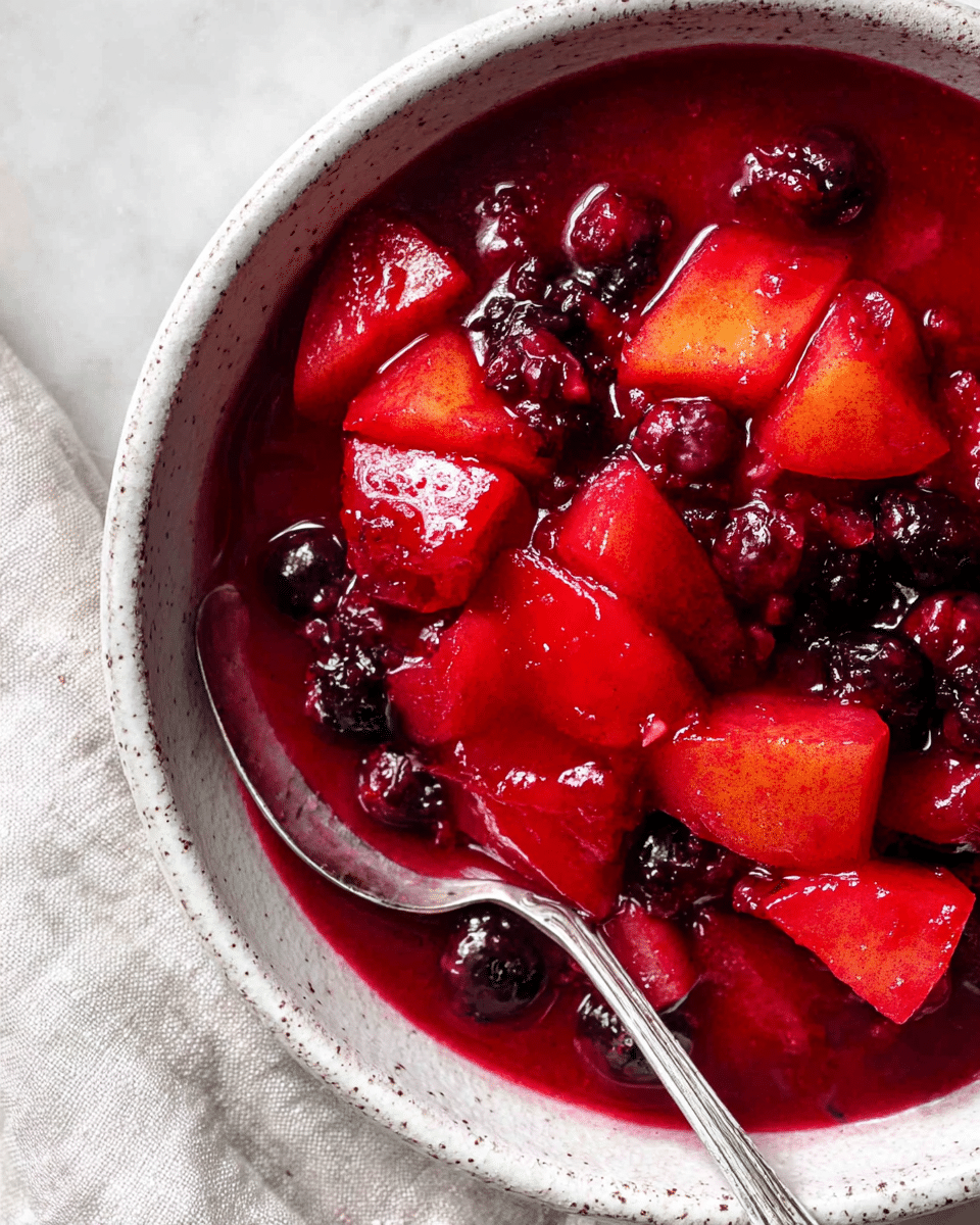A close-up view of a bowl filled with a chunky fruit compote that has three main layers: large, shiny, red apple pieces with a smooth texture and small dark red and purple berries that look juicy, all sitting in a thick, deep red syrup. The bowl is off-white with a speckled pattern around the rim and has a silver spoon partially visible inside. The background is a white marbled surface with a soft linen cloth next to the bowl. Photo taken with an iphone --ar 4:5 --v 7