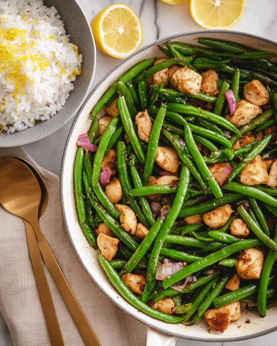 A close-up view of a white frying pan filled with bright green beans and golden-brown pieces of cooked chicken, mixed with small bits of red onions. The green beans are glossy, fresh, and evenly spread among the chicken pieces, which have a slightly crispy texture on the outside. To the right, there is a gray bowl holding white rice with some yellow lemon zest on top, and next to it are two lemon halves showing their juicy interior. A pair of golden-colored fork and spoon rest on a beige cloth napkin beside the pan. The whole scene is set on a white marbled surface. photo taken with an iphone --ar 4:5 --v 7