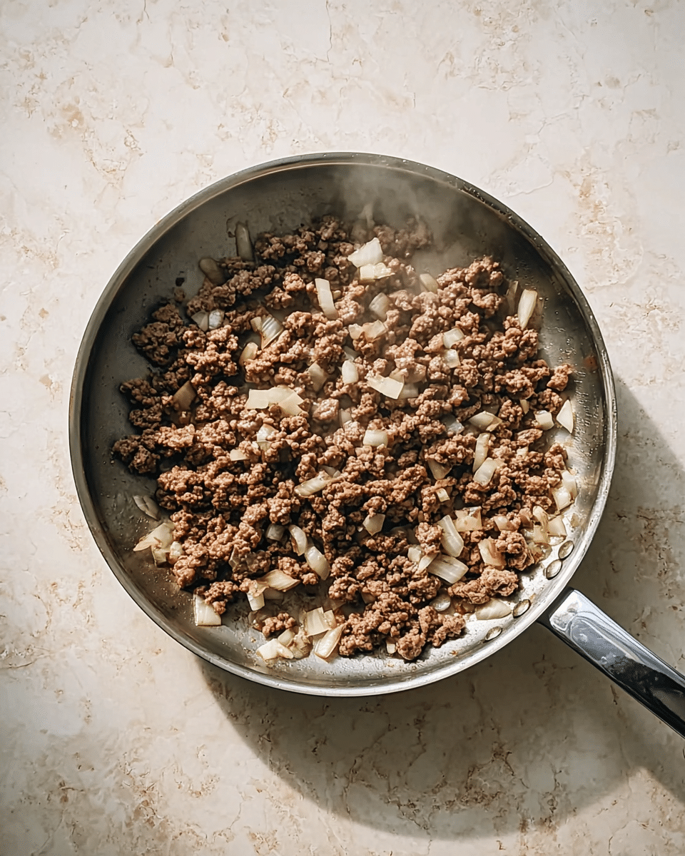 A frying pan with a silver handle holds a mix of brown cooked ground meat and small white onion pieces. The meat is broken into small chunks spread evenly in the pan, and some steam rises from the hot food. The pan sits on a surface with a white marbled texture that catches soft light, creating a warm and homely feel. photo taken with an iphone --ar 4:5 --v 7