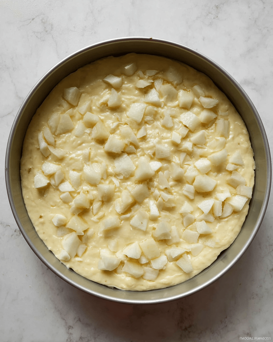 A round, silver baking pan is filled with one thick layer of pale yellow batter mixed with small, uneven chunks of white fruit scattered throughout the top. The batter looks smooth and creamy, with the fruit pieces adding texture and interest across the surface. The pan is placed on a white marbled surface. photo taken with an iphone --ar 4:5 --v 7