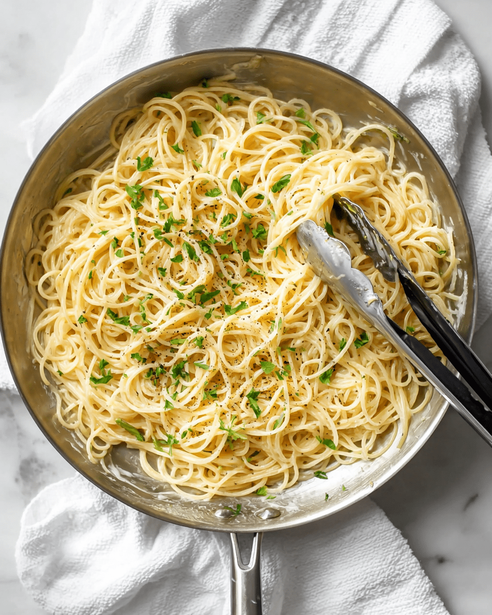 A round silver pan filled with creamy spaghetti noodles that have a light yellow color, coated evenly in sauce, scattered with small green parsley leaves and light black pepper flakes; silver tongs with black handles rest on the right side, partially lifting some noodles; the pan sits on a white cloth over a white marbled surface. photo taken with an iphone --ar 4:5 --v 7