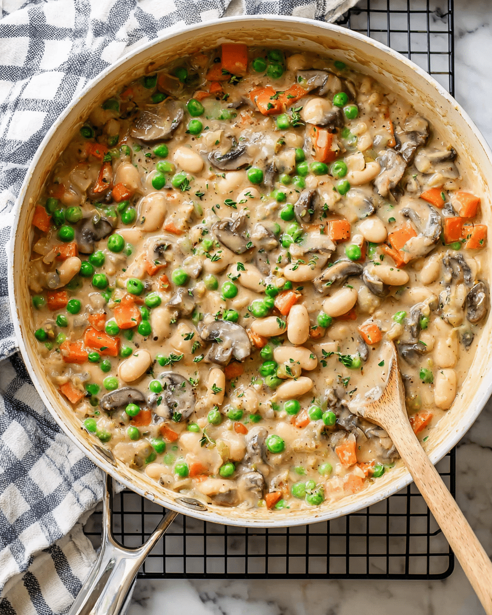 A round white pan filled with a creamy mix of layers: light beige sauce as the base, with white beans scattered throughout, bright green peas, small orange carrot cubes, and brown sliced mushrooms layered evenly. The creamy texture coats the beans and vegetables, with bits of green herbs sprinkled on top. A wooden spoon rests inside the pan on the right side, partly dipped into the mix. The pan handle is metallic, and the background shows a white marbled surface with a checked white and navy cloth and a black cooling rack beneath the pan. Photo taken with an iphone --ar 4:5 --v 7