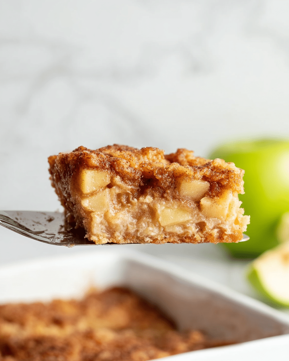 A single piece of golden-brown apple dessert held above a white dish by a metal spatula, showing a slightly crispy top layer with small apple chunks, a moist, creamy middle layer with a mix of light beige and cinnamon brown colors, and a slightly denser bottom layer matching the middle in texture; the background is a white marbled texture with a blurred green apple on the right and a small pale slice on the left, photo taken with an iphone --ar 4:5 --v 7