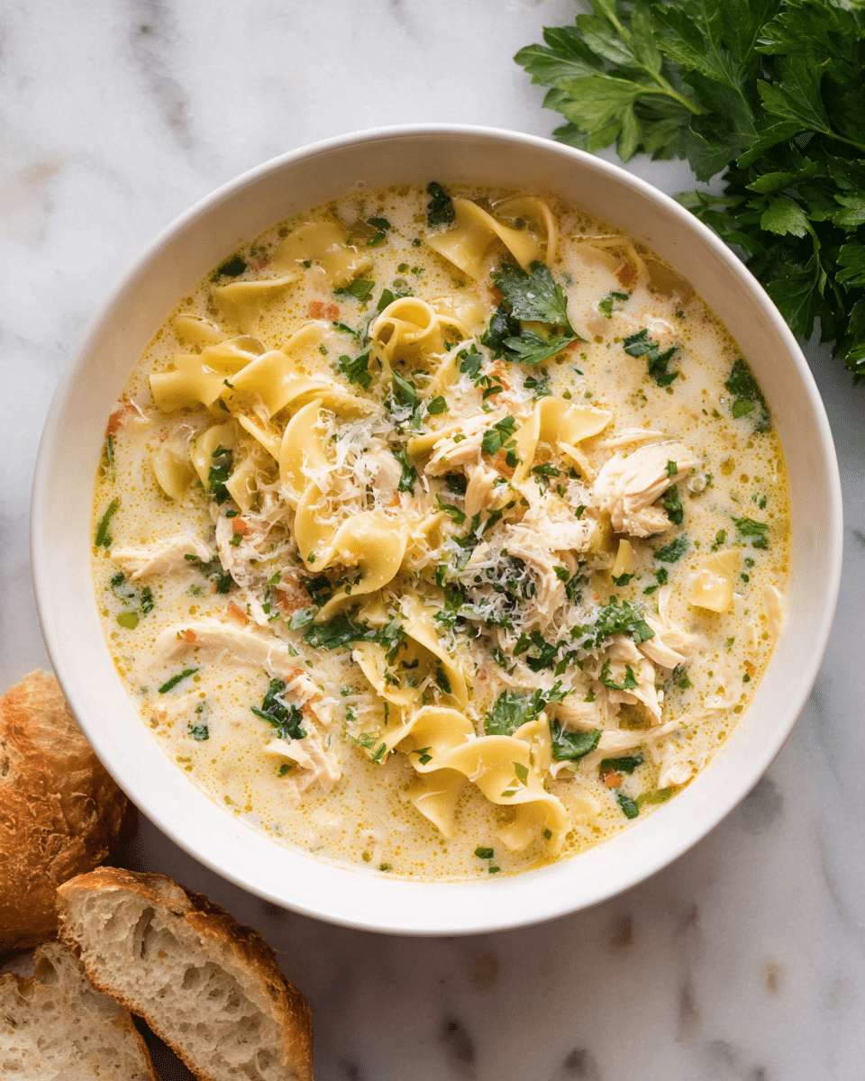 A white bowl filled with creamy chicken noodle soup with about three layers: the base is a thick light yellow creamy broth, floating on top are curly-edged yellow noodles, shredded light beige chicken pieces, and small chopped green leafy herbs scattered throughout, with some grated white cheese sprinkled on top. The bowl sits on a white marbled surface, with fresh green parsley on the top right corner and slices of crusty bread on the lower left corner, photo taken with an iphone --ar 4:5 --v 7