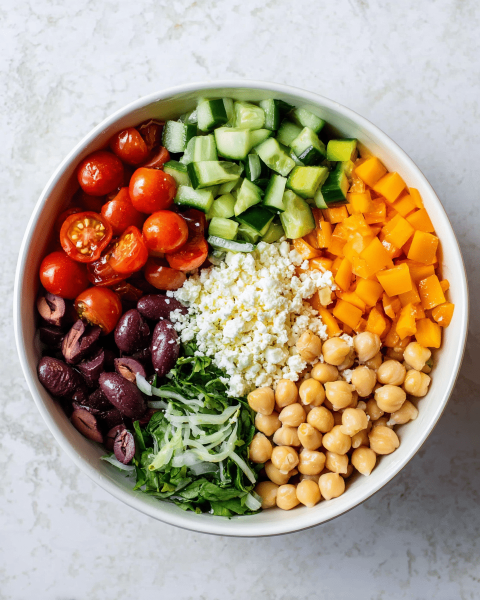 A white bowl is filled with six distinct layers of food arranged side by side in a circular pattern. Starting from the top left, there are bright red halved cherry tomatoes with a shiny texture. Next, there is a layer of chopped green cucumber pieces with dark skins. The top right section has small diced orange bell pepper pieces. Below that, on the bottom right, there is a pale beige layer of round chickpeas. To the left of the chickpeas, there is a pile of thinly sliced dark green leafy herbs. Finally, to the left of the herbs, there are dark purple olives with a smooth, shiny surface. In the center of the bowl, there is a pile of crumbled white cheese. The bowl sits on a white marbled textured surface. photo taken with an iphone --ar 4:5 --v 7