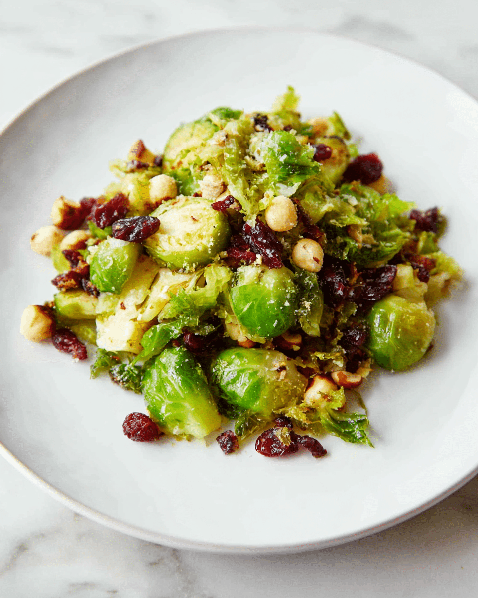 The dish shows a small pile of cooked Brussels sprouts pieces in shades of vivid green mixed with toasted hazelnuts in light brown and small pieces of dark red cranberries scattered throughout. The Brussels sprouts are slightly browned and have a rough texture, giving a sense of a light roasting or sauté. The ingredients sit on a plain white plate placed on a white marbled background. The overall look is fresh and healthy with a mix of colors from the Brussels sprouts, nuts, and cranberries. photo taken with an iphone --ar 4:5 --v 7