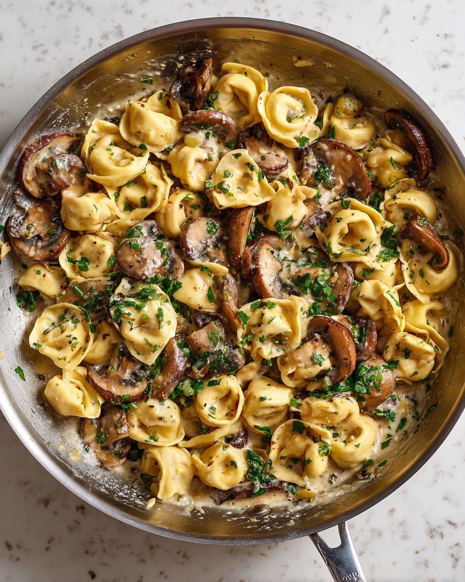 The image shows a metal pan filled with a creamy pasta dish featuring two types of pasta shapes: round tortellini and small folded tortellini, both in a light golden color. Mixed throughout are browned slices of mushrooms adding a rich dark brown contrast. The creamy sauce has a smooth, glossy texture and light beige color that coats the pasta and mushrooms evenly. The dish is sprinkled with fresh green chopped herbs, giving a fresh and colorful touch. The pan rests on a white marbled surface. Photo taken with an iphone --ar 4:5 --v 7