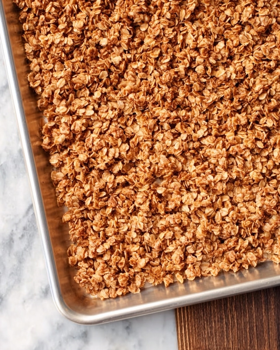 A close-up shot of a silver baking tray filled with even layers of golden brown granola spread out flat. The granola has a rough, crumbly texture with visible oats and small clusters, covering the entire tray surface. The tray lies on a white marbled textured surface, emphasizing the warm colors of the granola. photo taken with an iphone --ar 4:5 --v 7