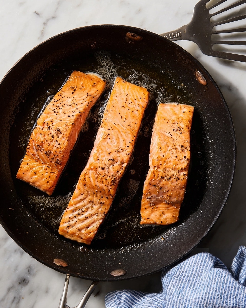 Three pieces of cooked salmon fillets lie in a black skillet. Each fillet is long and rectangular with a light orange color, showing grill marks and sprinkled with black pepper. The skillet has traces of oil and small bubbles around the fish. It rests on a white marbled surface, and to the right side, there is a metal spatula. A blue and white striped cloth is partially visible at the bottom right corner. photo taken with an iphone --ar 4:5 --v 7