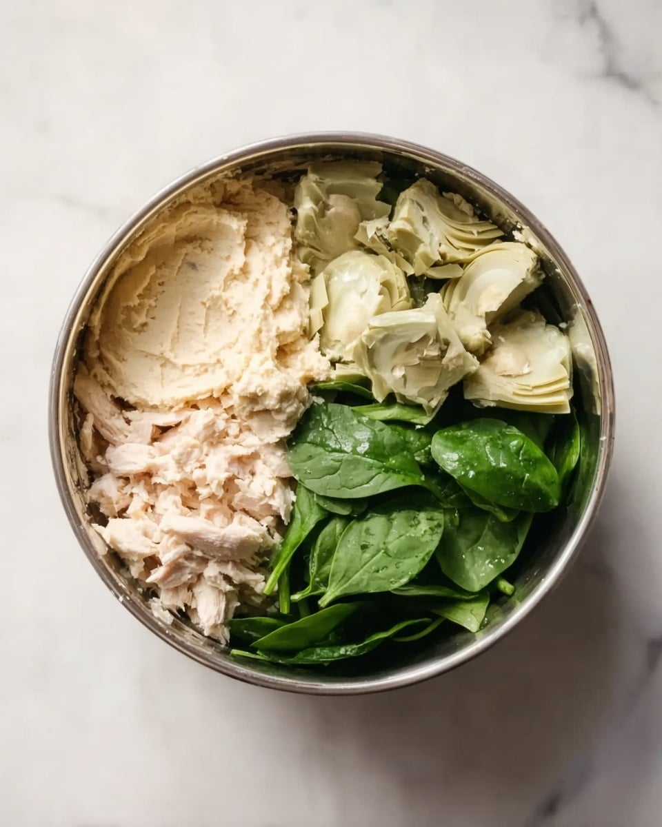 A round metal bowl sits on a white marbled surface, filled with four distinct layers of ingredients arranged side by side. On the left, there is a light beige layer of soft, spreadable mixture. Next to it are pale green pieces of chopped artichokes with a slightly rough texture. Below the artichokes, fresh, bright green spinach leaves are piled, smooth and leafy. On the far left, small, slightly shredded pieces of light pinkish white cooked chicken rest against the mixture. The ingredients create a colorful, fresh combination in the bowl. Photo taken with an iphone --ar 4:5 --v 7