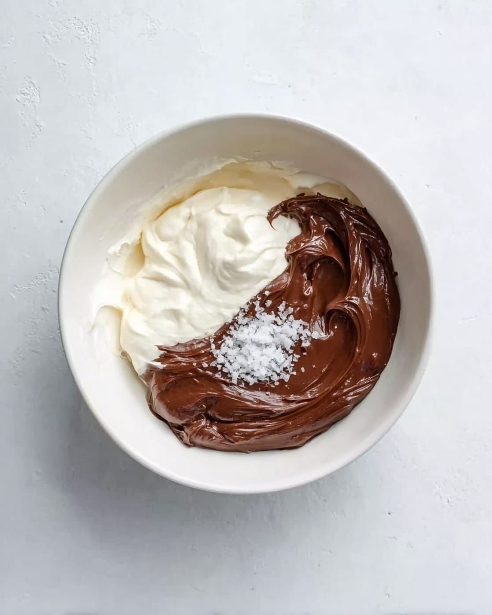 A white bowl contains three main layers of ingredients. On the left side, there is a thick, creamy white layer. Next to it on the right is a smooth, rich dark brown chocolate layer. In the middle, on top of the chocolate, there is a small pile of fine white salt crystals. The bowl sits on a white marbled surface. Photo taken with an iphone --ar 4:5 --v 7