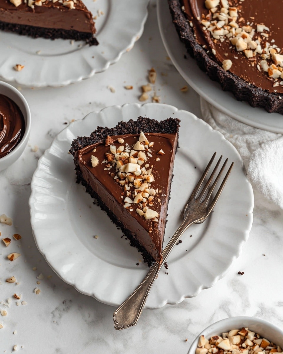 A slice of chocolate tart is placed on a white scalloped-edge plate sitting on a white marbled surface. The tart has three layers: a dark crumbly crust at the bottom, a smooth and shiny milk chocolate filling in the middle, and is topped with small pieces of chopped light brown nuts scattered on top. Next to the tart slice lies a vintage silver fork. Around this plate, parts of other white scalloped-edge plates with similar chocolate tart slices topped with nuts and a bowl with chocolate filling are visible. Photo taken with an iphone --ar 4:5 --v 7