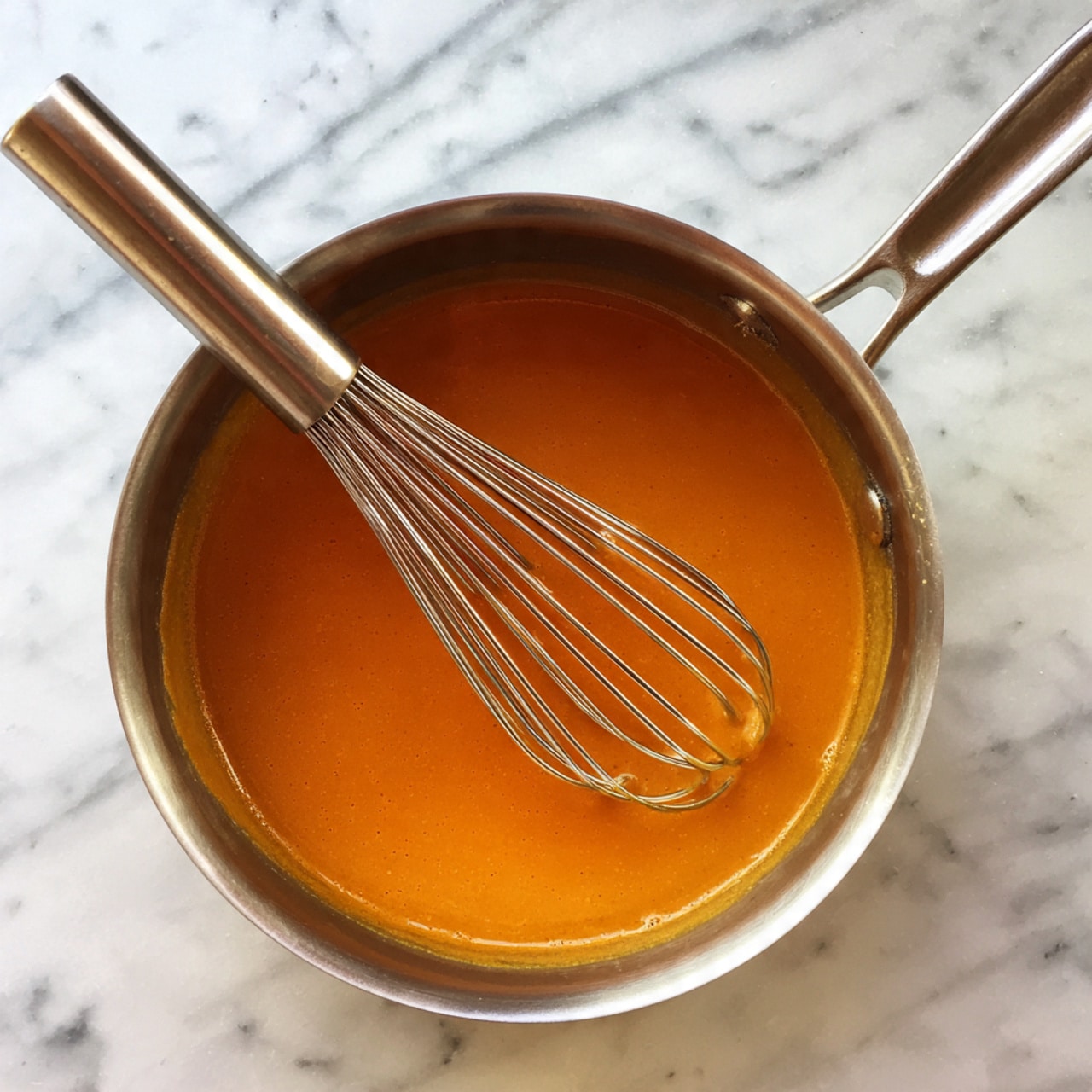A small silver pan filled with a smooth, deep orange sauce that has a shiny texture. A silver whisk is resting inside the pan, partially covered by the sauce, with its handle pointing toward the upper left corner of the image. The pan is sitting on a white marbled surface. photo taken with an iphone --ar 4:5 --v 7