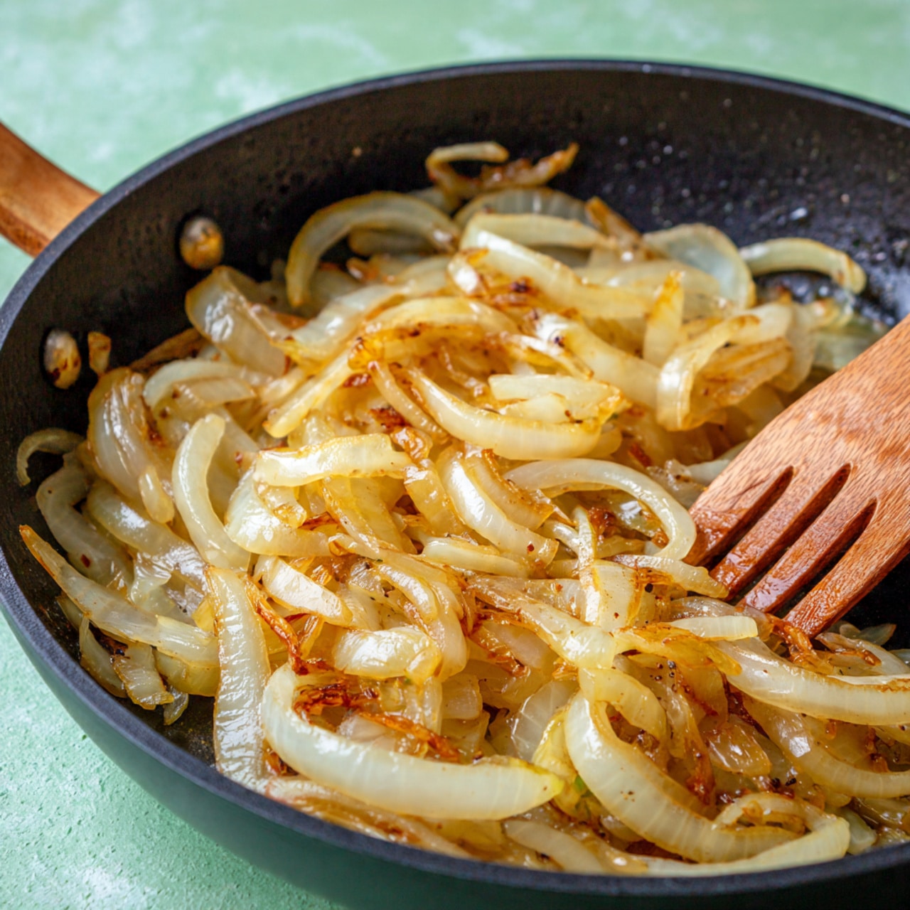 A close-up of a black pan filled with soft cooked onions that are golden brown with some caramelized parts. The onions have a shiny, slightly oily texture and are cut into thin strips, layered loosely in the pan. A wooden fork is stirring the onions on the right side, with the fork partially buried in the onion layers. The background shows a soft, blurred surface with light green tones. Photo taken with an iphone --ar 4:5 --v 7
