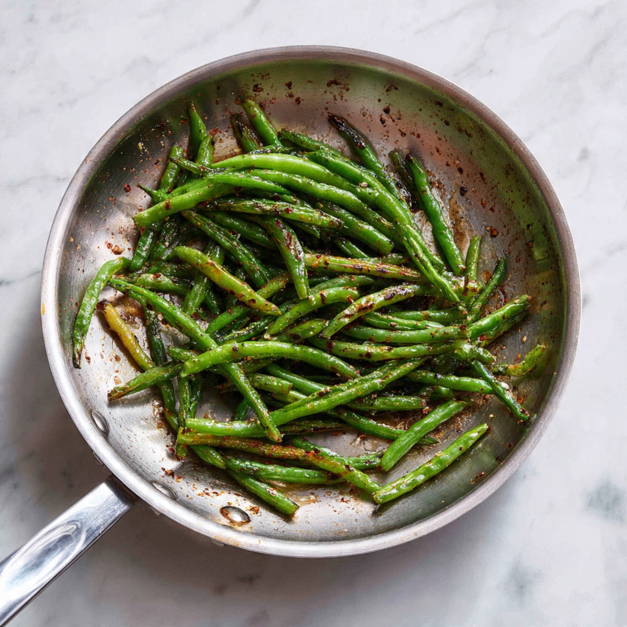 The image shows a silver frying pan placed on a white marbled surface, containing a single layer of cooked green beans. The green beans are slightly browned and speckled with grill marks, scattered unevenly across the pan’s smooth, shiny interior. The pan’s metal handle extends out to the left, and the background remains clean with the white marbled texture clearly visible. photo taken with an iphone --ar 4:5 --v 7