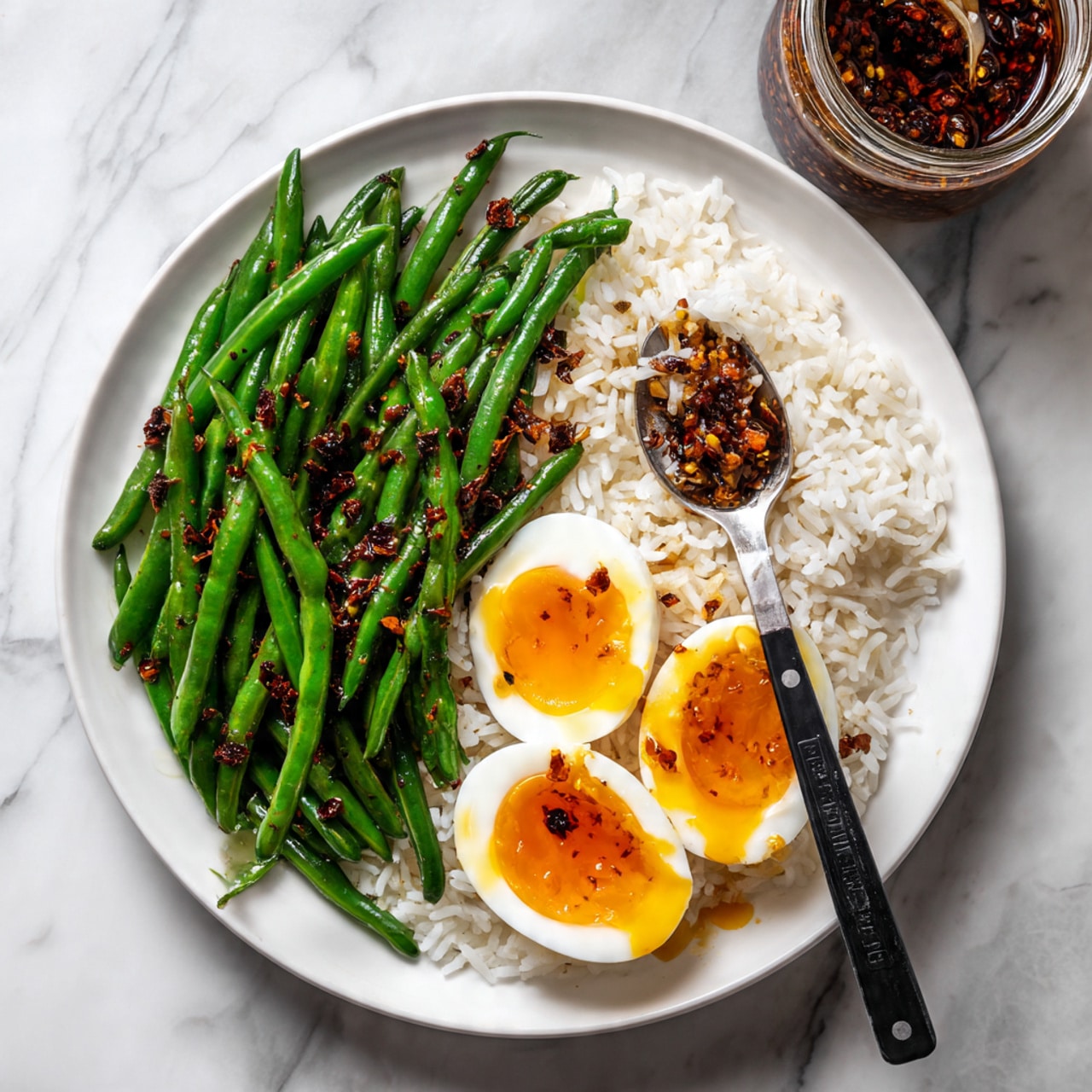 A white plate on a white marbled surface holds three main layers: on the left side, bright green cooked green beans with a slightly shiny texture and small bits of dark red chili flakes scattered on them; on the right side, fluffy white rice with a few specks of chili flakes over it; on top of the rice, two halves of a soft-boiled egg with a bright yellow yolk that is slightly runny, one half cut further with the yolk flowing out onto the rice. A black fork rests on the rice, touching the cut egg yolk. In the top right corner above the plate, there is a jar containing dark chili oil and a spoon. photo taken with an iphone --ar 4:5 --v 7