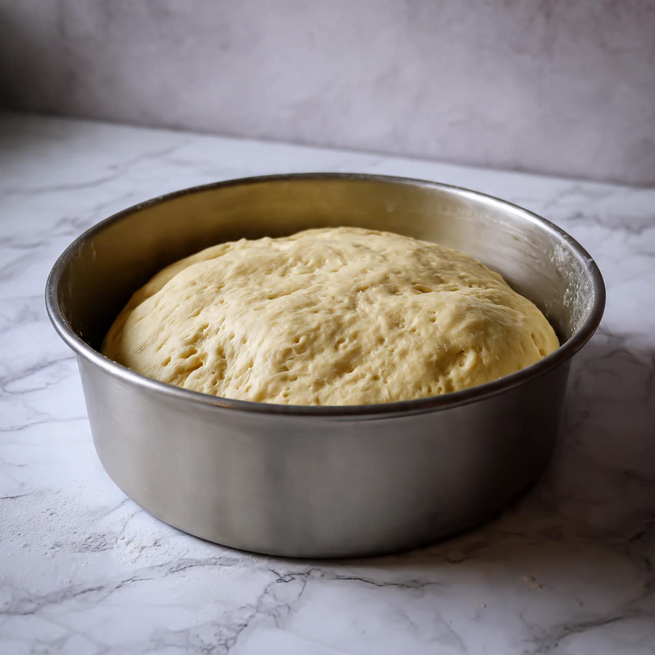 A light beige dough sits in a smooth silver metal bowl, puffed up as it has risen, showing small air bubbles on its surface. The dough has a soft and slightly shiny texture, gently filling the round bowl which has a metallic sheen inside and out. The background is a white marbled texture, giving a clean and bright look to the scene. photo taken with an iphone --ar 4:5 --v 7