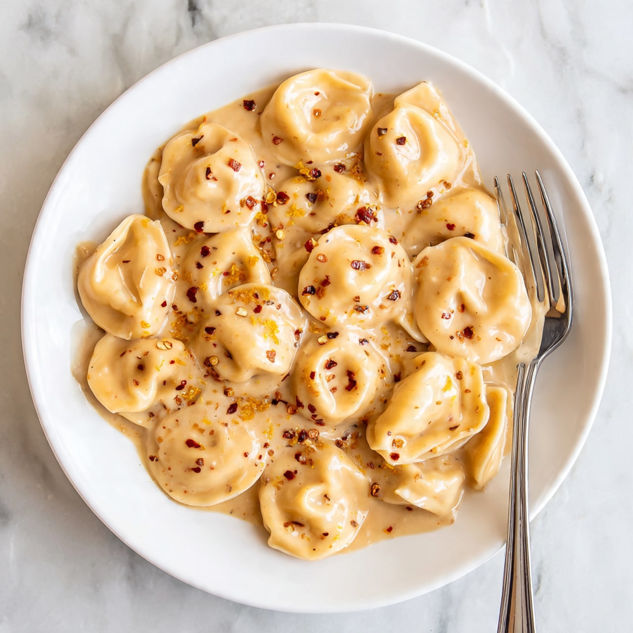 A white round plate holds one layer of round tortellini pasta, each piece light beige and covered in creamy, smooth beige sauce with specks of black pepper. The tortellini are scattered with small red chili flakes and fine yellow zest on the sauce. A silver fork is placed on the right side on the plate, partly under the pasta. The background is a white marbled texture. Photo taken with an iphone --ar 4:5 --v 7