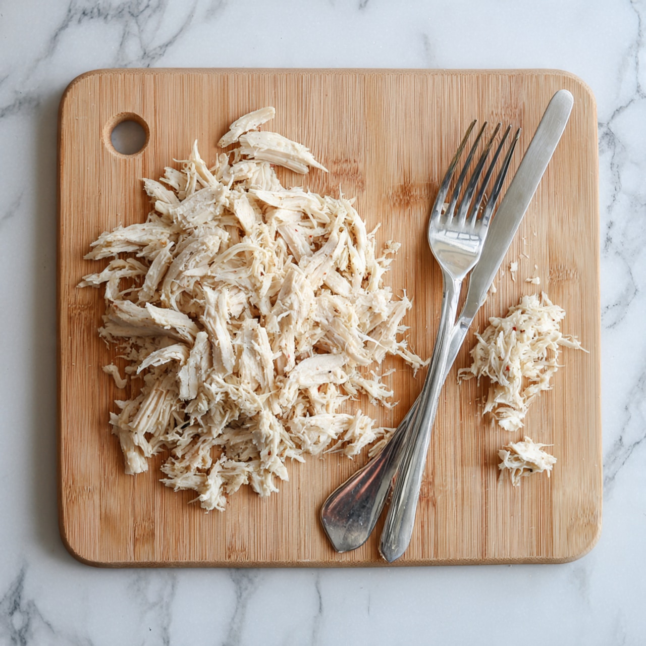 A light brown wooden cutting board sits on a white marbled surface. On the board, there is a pile of shredded cooked chicken in pale beige and off-white colors. Two silver forks are placed on the right side of the chicken with some shredded pieces on them, showing the texture of the pulled chicken. The overall look is simple, fresh, and ready for use in a recipe. photo taken with an iphone --ar 4:5 --v 7