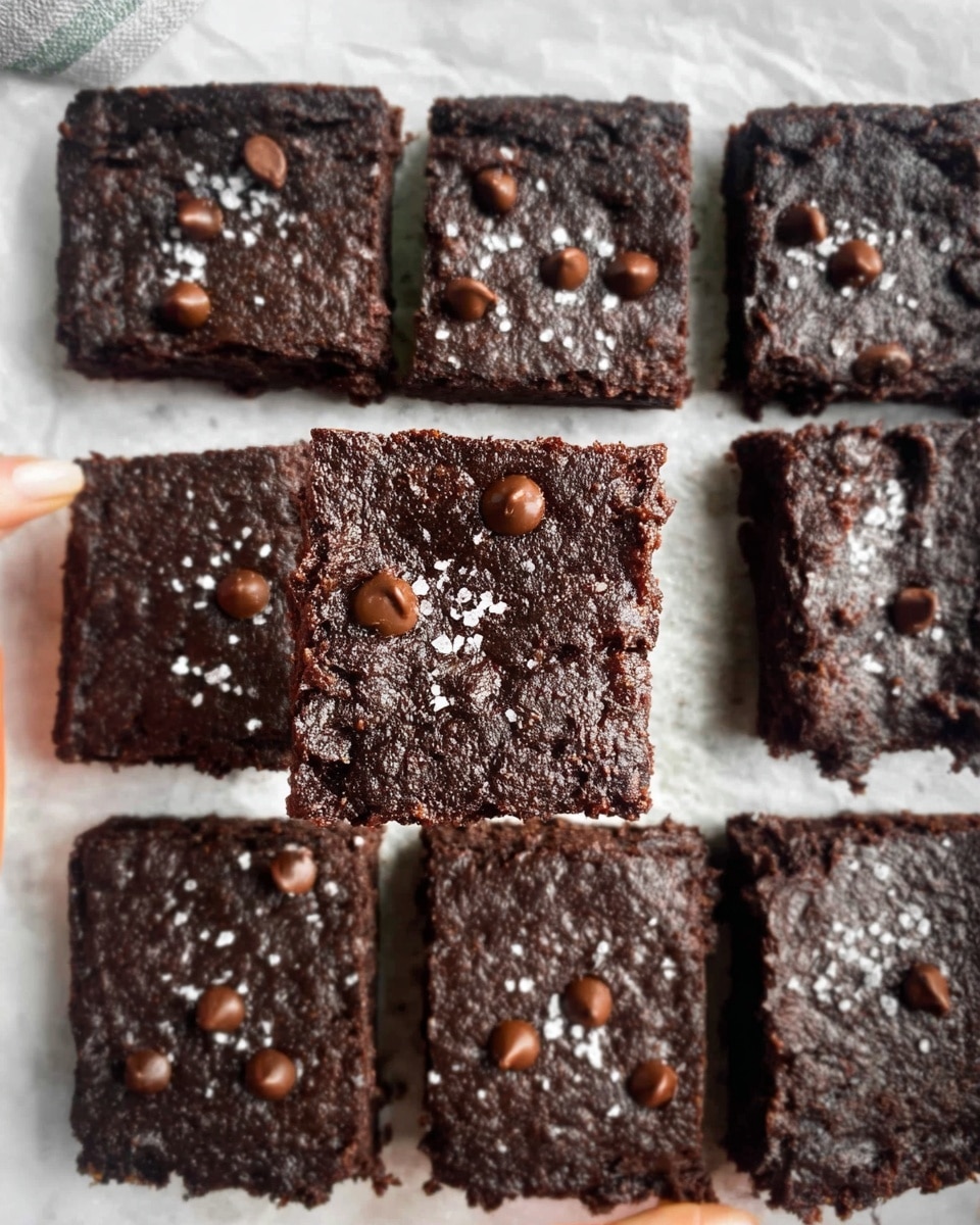 The image shows nine square brownies arranged in a 3x3 grid on white parchment paper over a white marbled surface. Each brownie is dark brown with a soft, moist texture and contains chocolate chips scattered on top, adding small round spots of lighter brown. The top of the brownies is sprinkled lightly with coarse white sea salt. One brownie in the center is held up by a woman's hand, showing its thick single layer, revealing a rich and dense chocolate inside. The edges of the brownies appear slightly crumbly. Photo taken with an iphone --ar 4:5 --v 7