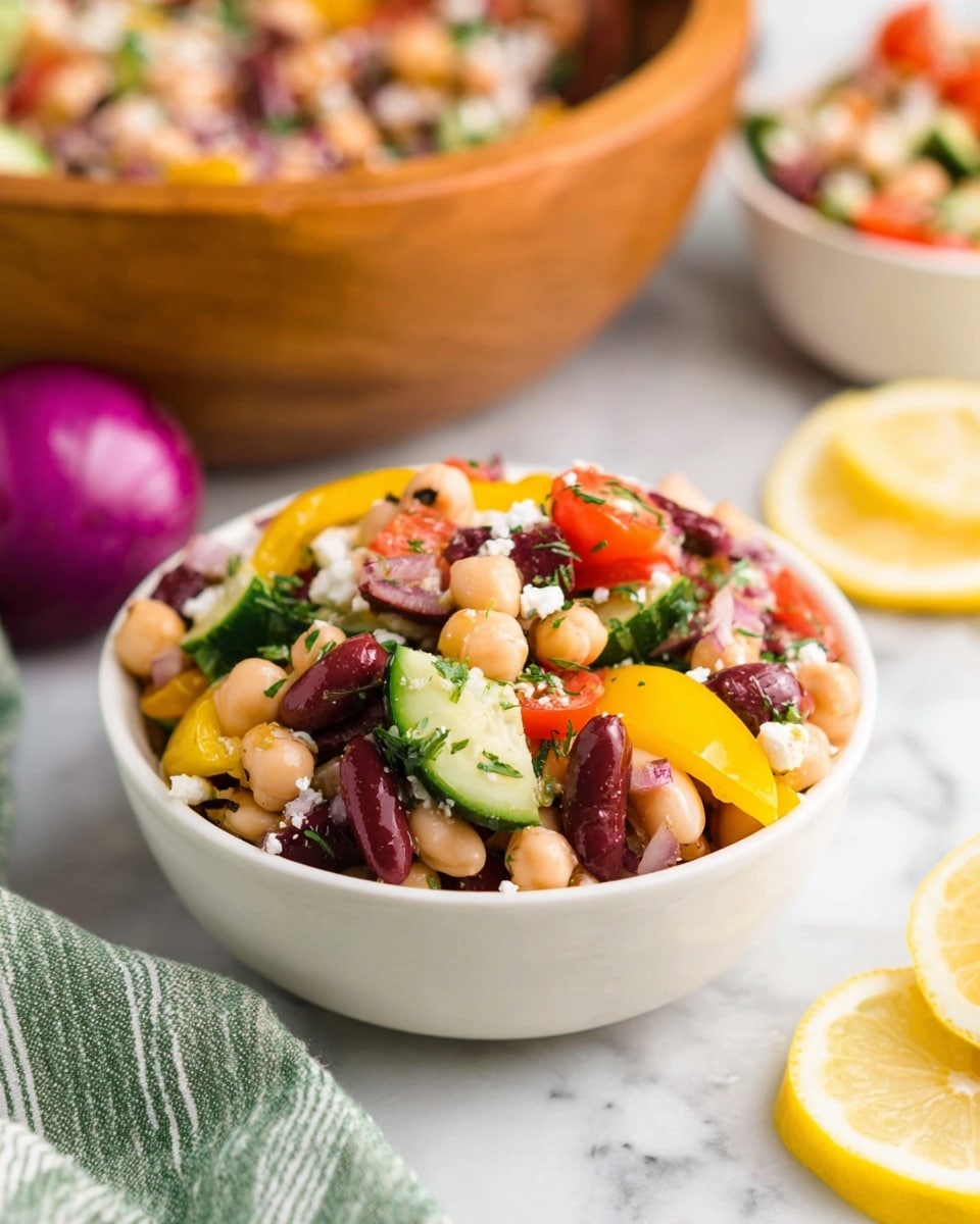 A small white bowl filled with a colorful bean salad showing three main layers: the base layer of white beans, kidney beans, and chickpeas in soft beige, dark red, and light tan; the middle layer of bright yellow bell peppers and sliced green cucumber with a fresh texture; and the top layer of halved red cherry tomatoes, sliced dark purple olives, small bits of white cheese, and finely chopped green herbs. The bowl sits on a white marbled surface with lemon halves and a piece of red onion in the background, next to a green and white striped cloth. A larger wooden bowl with more of the salad is blurred in the background. Photo taken with an iphone --ar 4:5 --v 7