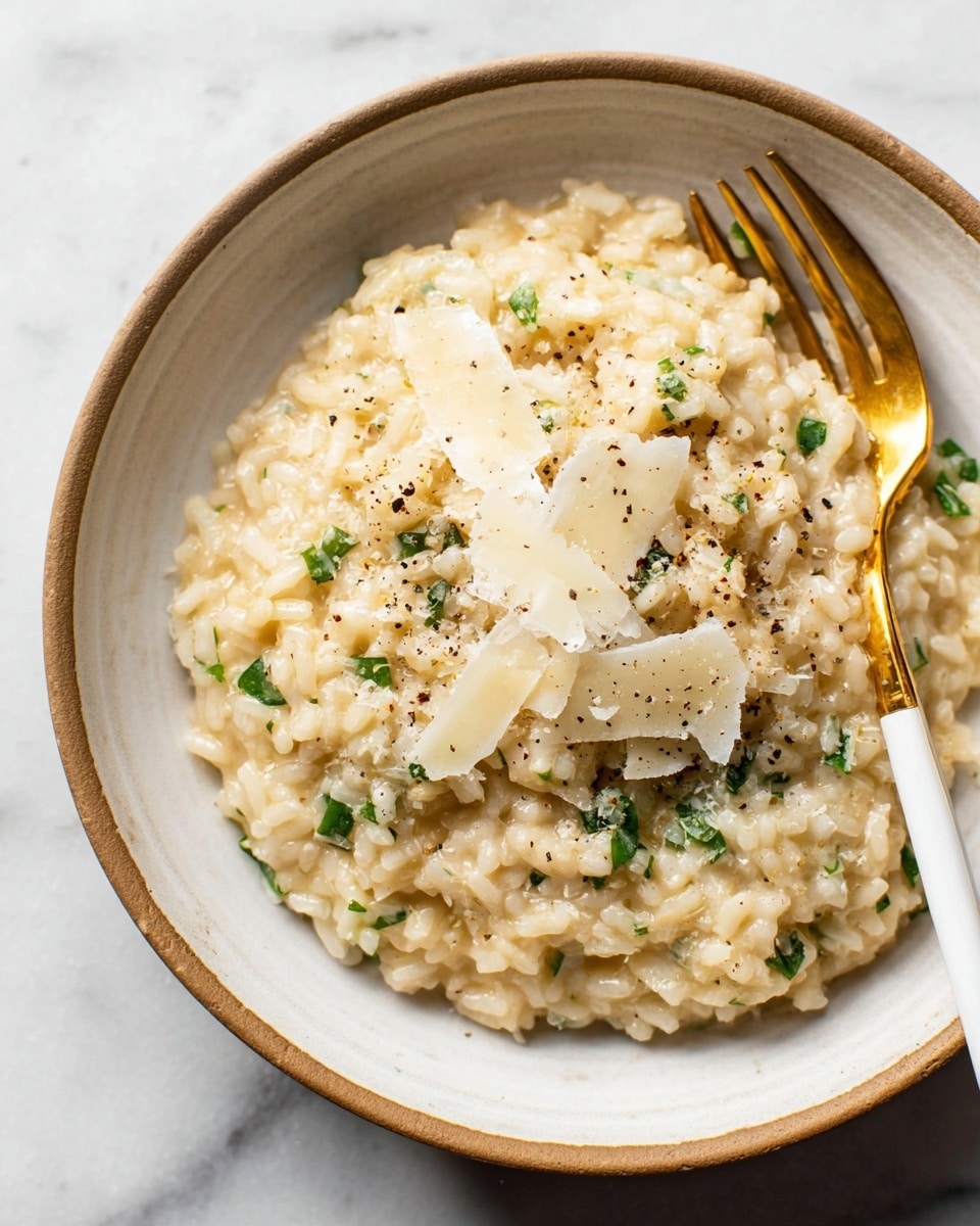 A close-up view of creamy risotto served in a white bowl with a light brown rim. The risotto is light beige with a soft, slightly sticky texture, sprinkled with small green herb pieces and topped with thin, white cheese slices. Black pepper specks are scattered on top, adding contrast. A gold fork with a white handle rests on the side of the bowl, partially buried in the risotto. The bowl is set on a white marbled surface. photo taken with an iphone --ar 4:5 --v 7
