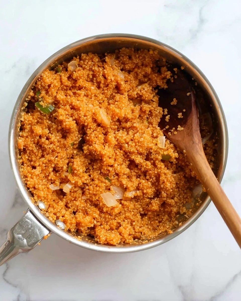 The image shows a stainless steel pan filled with cooked quinoa that is fluffy and has a warm orange color, mixed with small pieces of translucent onions and some soft green vegetable bits, with a wooden spoon resting inside the pan on the right side. The pan is placed on a white marbled surface that adds a clean and bright look to the scene. Photo taken with an iphone --ar 4:5 --v 7
