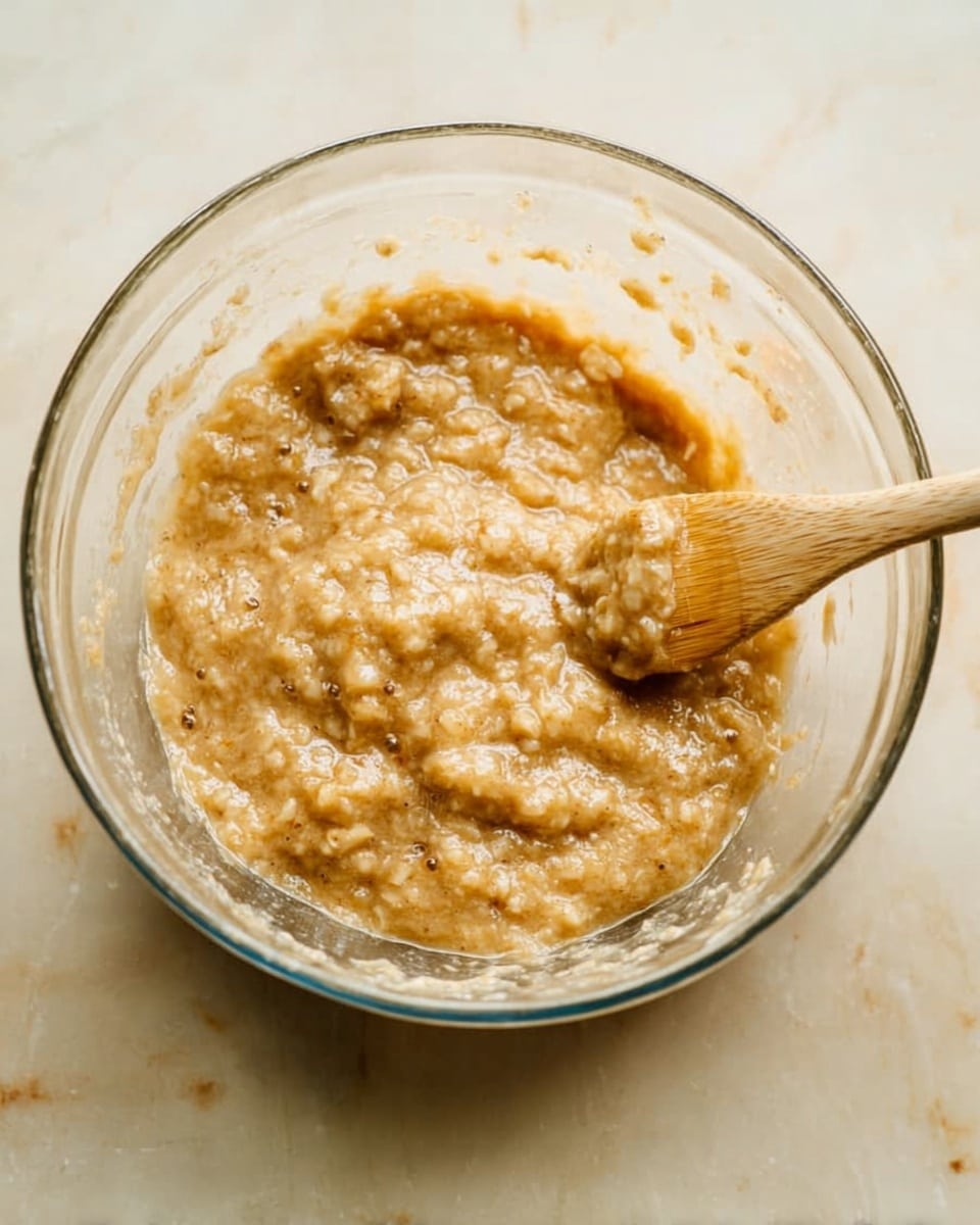 A clear glass bowl sits on a white marbled surface, filled with mashed banana mix that is light brown with some small chunks and bubbles, giving it a slightly textured look. A light wooden spatula rests inside the bowl, partially covered by the banana mixture. The glass bowl is round and transparent, showing the thick, wet banana mash inside. The background and surface are smooth white marble, adding a clean and simple feel to the image. Photo taken with an iphone --ar 4:5 --v 7