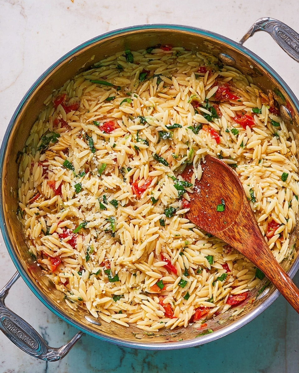 A large shallow silver pan is filled with cooked orzo pasta mixed with bits of red tomatoes and green herbs, likely parsley or spinach, evenly spread across. A wooden spoon is placed inside the pan stirring the orzo, showing the shiny texture of the noodles mixed with small flecks of white cheese sprinkled on top. The pan rests on a white marbled surface, with both of its side handles visible and sparkling clean, giving a fresh and simple look to the dish. Photo taken with an iphone --ar 4:5 --v 7