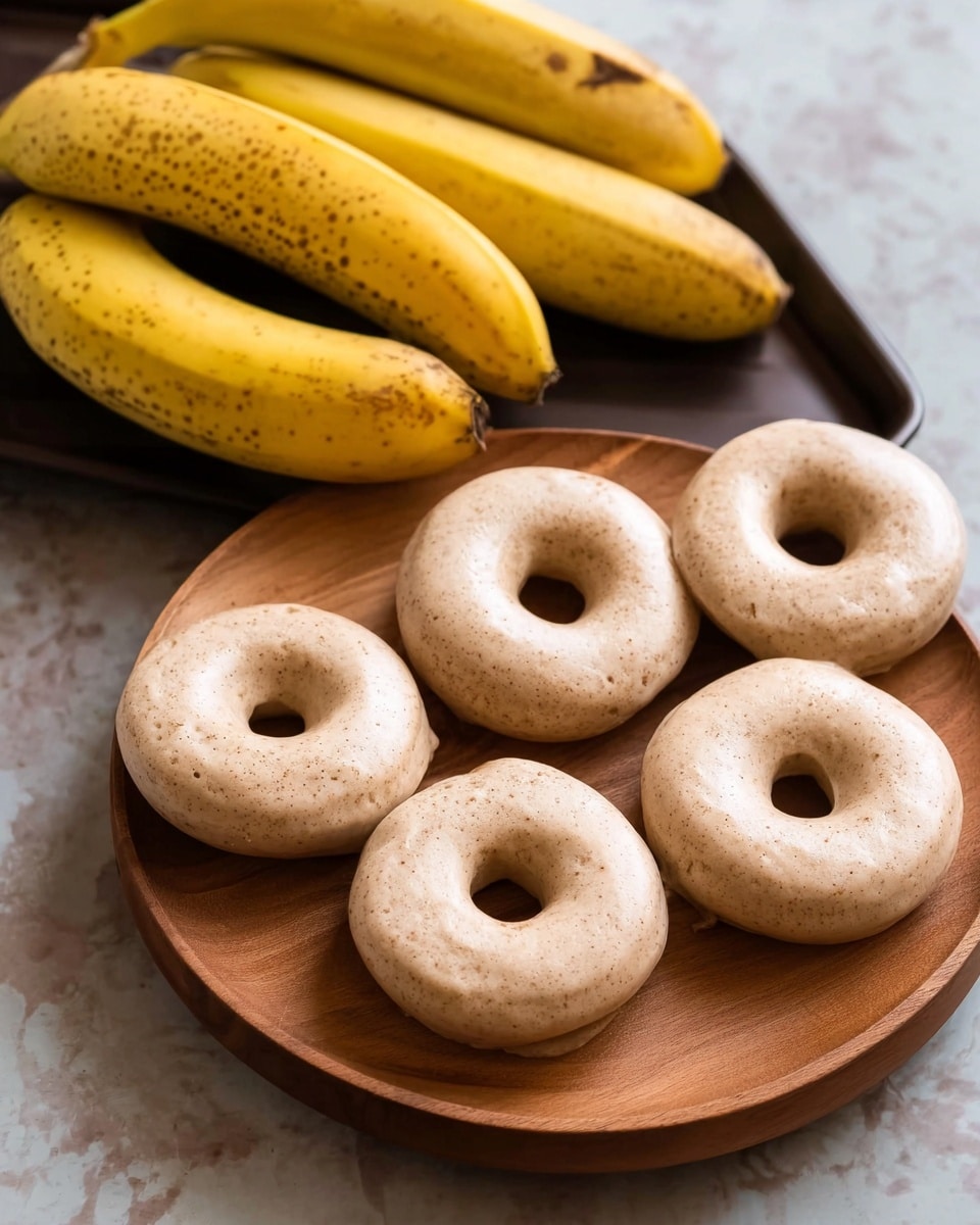 The image shows five raw doughnuts with a light tan color and visible tiny specks, arranged in a loose circle on a round wooden board. The doughnuts have a smooth, slightly shiny surface with a soft, thick texture and a central hole in each. Behind the board, there are three ripe yellow bananas with brown spots resting on a dark tray. The scene is set on a white marbled textured background. photo taken with an iphone --ar 4:5 --v 7