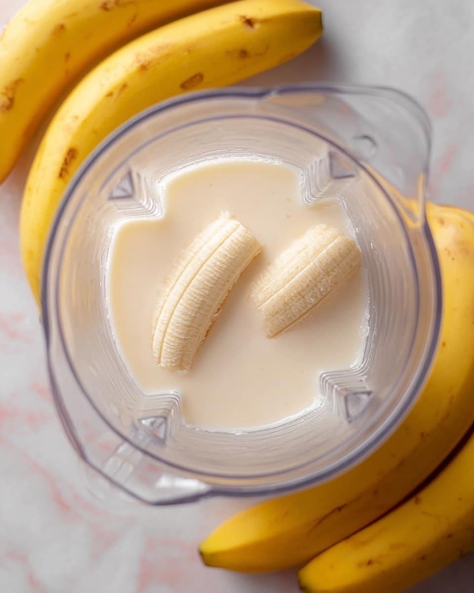 A clear blender container sits on a white marbled surface, holding two small peeled banana halves partially covered by a smooth, light cream liquid. A bunch of whole yellow bananas with some brown spots rests next to the blender, adding a touch of bright yellow to the scene. The image captures a close-up overhead view, focusing on the soft textures of the banana and the creamy liquid inside the container. Photo taken with an iphone --ar 4:5 --v 7