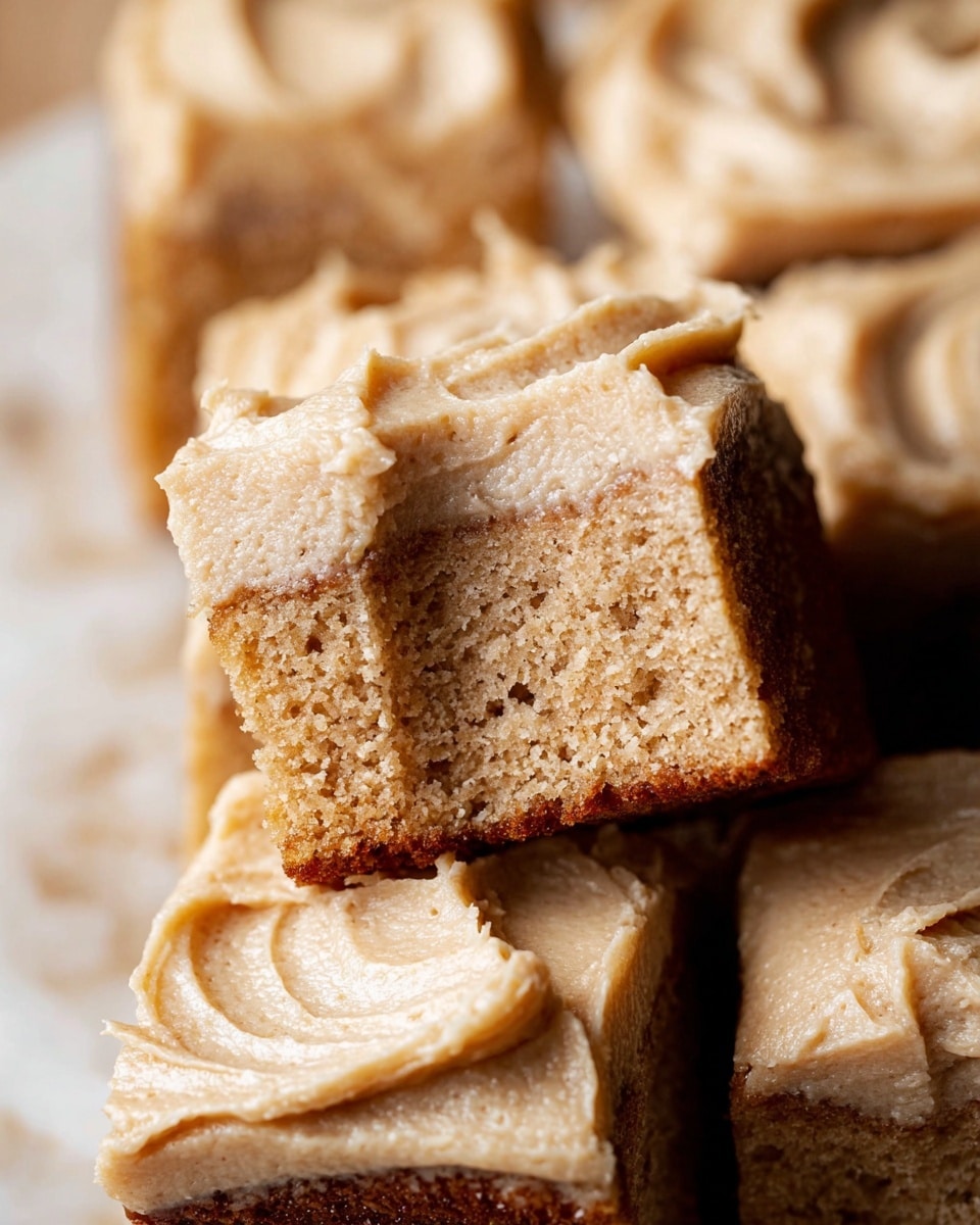 A close-up view of square cake pieces stacked on a white marbled surface, each piece having two layers: the bottom layer is a moist, light brown spongy cake with small air holes and a slightly rough texture, while the top layer is a thick, creamy light tan frosting with smooth swirls and spread marks. The frosting covers the cake evenly, with some raised and textured peaks showing its soft, fluffy consistency. The image focuses on one piece that is bitten into, revealing the soft cake inside with the creamy frosting on top. Photo taken with an iphone --ar 4:5 --v 7