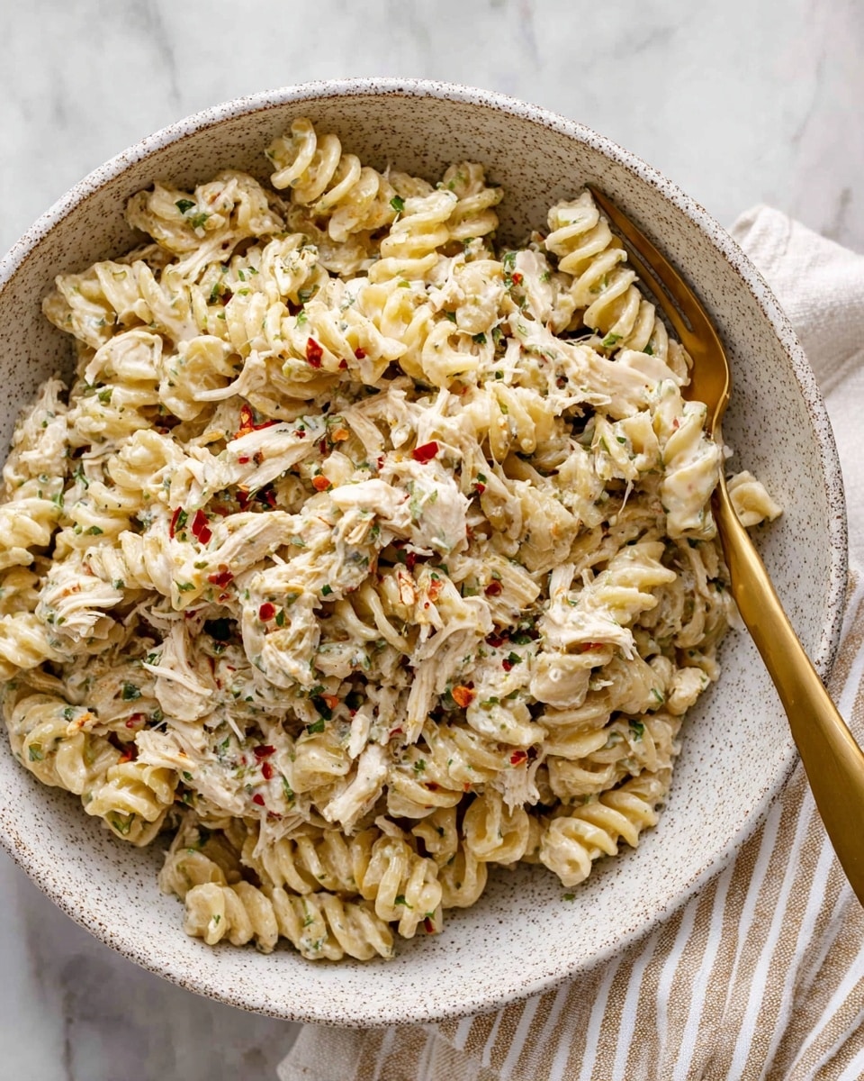 A large white speckled bowl filled with creamy pasta made of bite-sized twist noodles mixed with shredded white chicken. The pasta is coated in a thick, white sauce with visible small green herbs and red chili flakes scattered throughout, adding color and texture. The dish has a slightly rough texture from the shredded chicken and herbs. A fork with a golden handle rests inside the bowl, slightly lifting some pasta. The bowl sits on a white marbled surface with a beige and white striped cloth partially visible under the bowl. photo taken with an iphone --ar 4:5 --v 7
