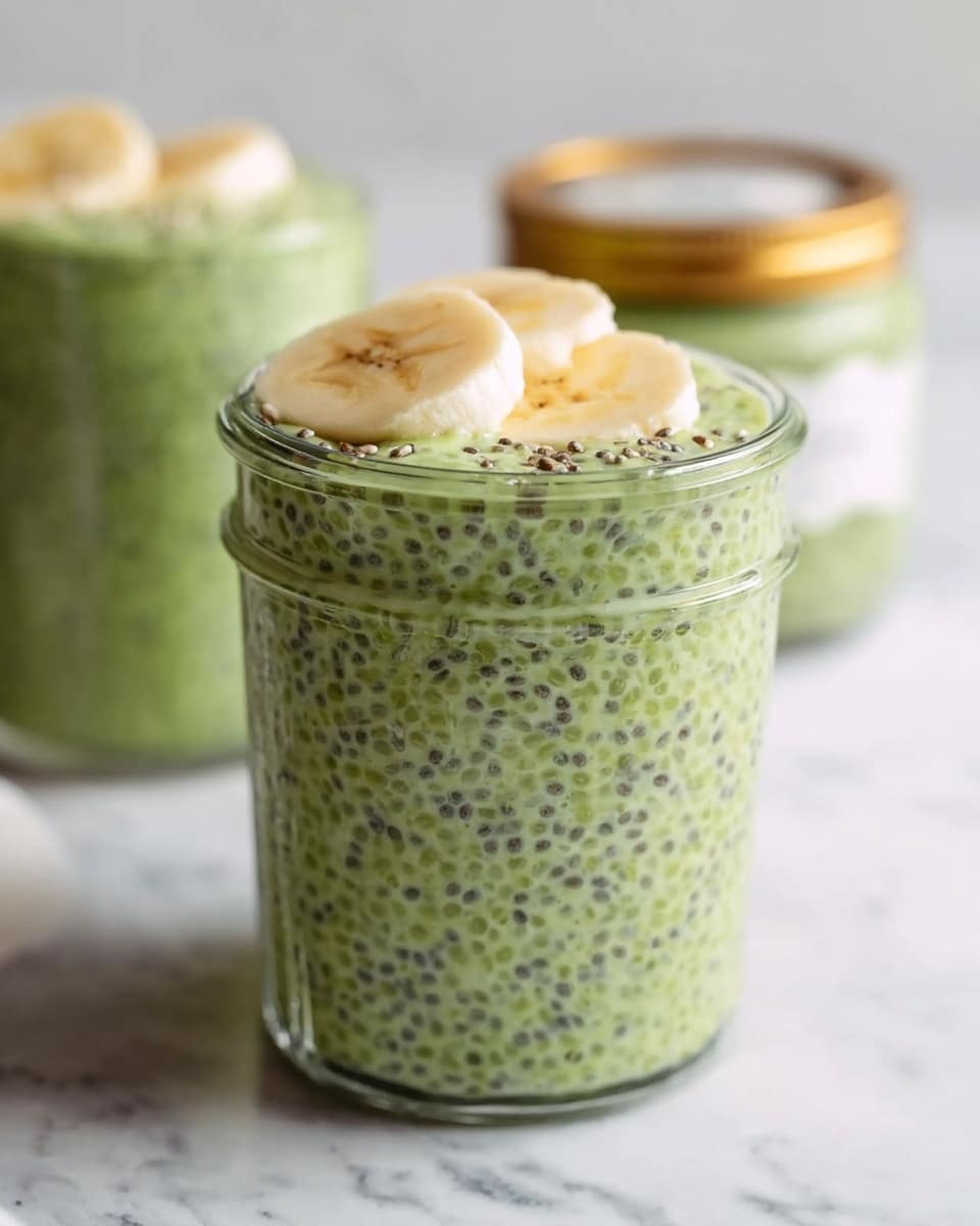 The image shows a clear glass jar filled with a thick, green chia pudding that has a textured mix of tiny black chia seeds evenly spread throughout the creamy green base. On top of the pudding, several slices of light yellow banana are placed partially inside the jar, adding a soft, curved layer. In the background, there is another similar jar slightly out of focus and a blurred jar with a label and a gold lid, all placed on a white marbled surface. The overall look is fresh and healthy with soft natural lighting. photo taken with an iphone --ar 4:5 --v 7