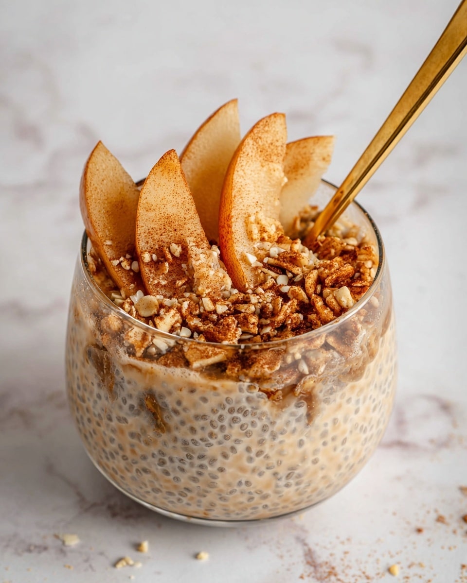 A close-up view of a clear glass filled with three main layers: the bottom layer is a creamy beige chia pudding with visible seeds creating a textured pattern; the middle layer consists of thin, soft apple slices with a light golden-brown color, slightly overlapping and curling around the edges; the top layer is a sprinkle of crushed nuts and cinnamon, adding a rough, crumbly texture in warm brown tones. A shiny gold spoon is inserted on the right side of the glass, partly covered by the layers. The background is a white marbled texture with small scattered bits of nuts. Photo taken with an iphone --ar 4:5 --v 7