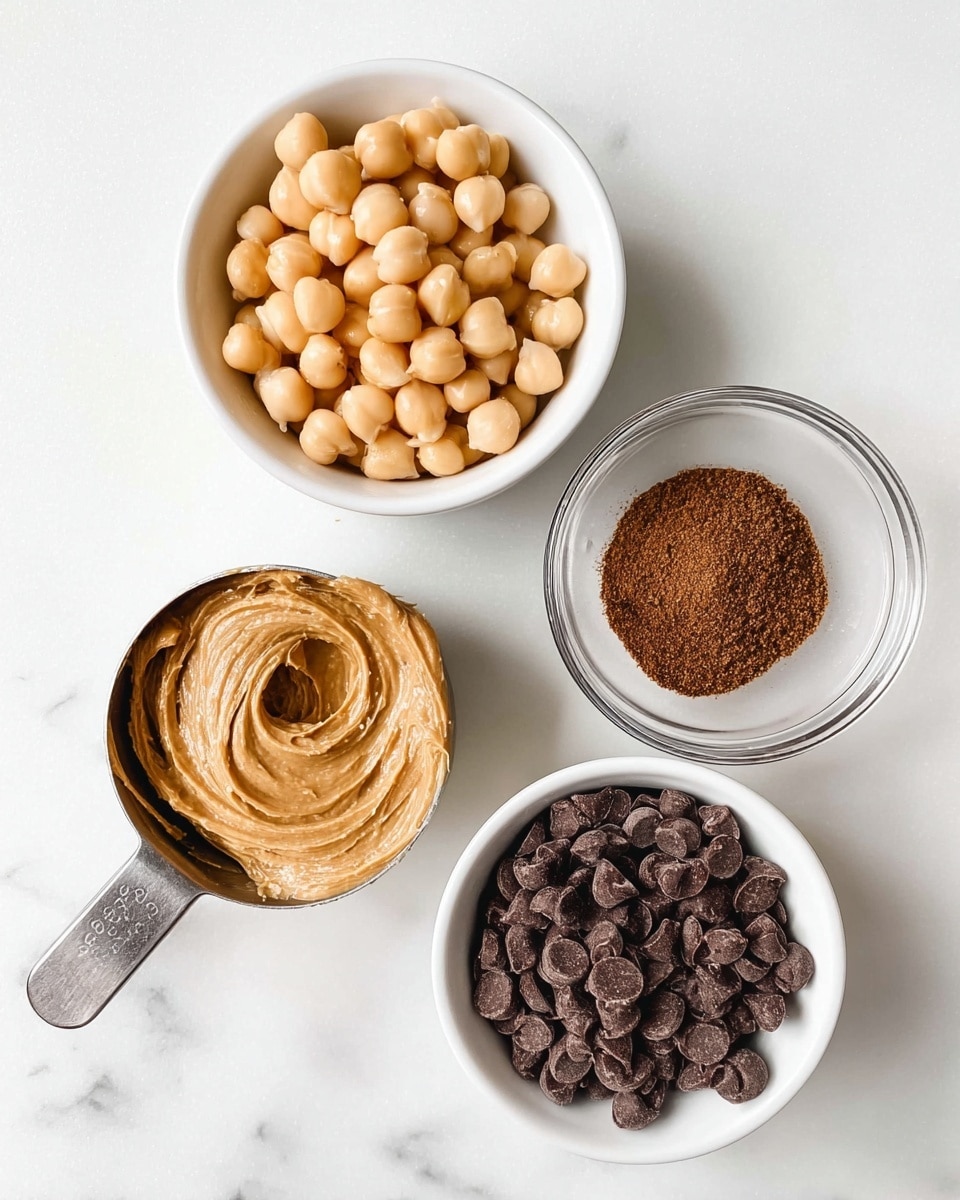 This image shows four containers of ingredients placed on a white marbled surface. At the top left, there is a white bowl filled with pale tan chickpeas, smooth and round. Below it, a metal measuring cup holds light brown creamy peanut butter with swirled texture. To the right of the peanut butter, a small clear glass bowl contains finely ground dark brown powder spices. At the bottom right, another white bowl is filled with dark brown chocolate chips, irregularly shaped and stacked loosely. The overall arrangement is clean and simple with a light, bright background. photo taken with an iphone --ar 4:5 --v 7