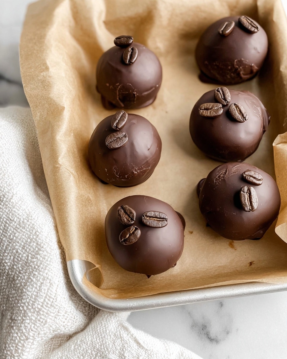 A tray lined with light brown parchment paper holds seven round chocolate balls, each coated smoothly with dark chocolate that shows light reflections and small texture marks. On top of each ball, there are three dark brown coffee beans arranged closely together. The tray is placed on a white marbled surface and a white textured cloth is partially visible on the left side of the image. photo taken with an iphone --ar 4:5 --v 7