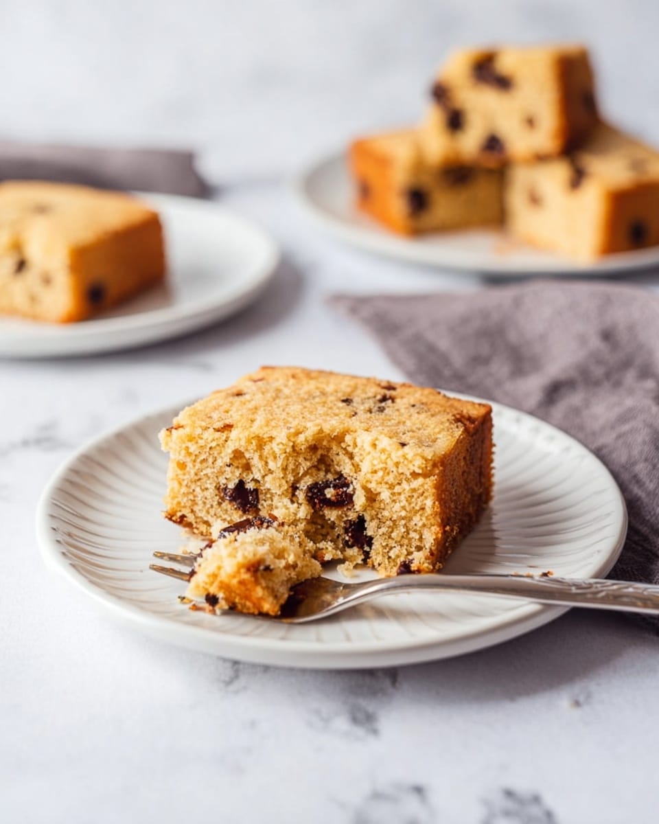 The image shows a soft, square piece of light brown cake with dark chocolate chips inside, placed on a white plate with a simple pattern around the edge. The cake is moist and crumbly, with one small portion broken off and resting on a fork next to the main piece. In the background, more pieces of the same cake are slightly blurred, lying on a white marbled surface. There is also a folded gray cloth and another white plate with a cake piece visible in the upper right corner. The overall look is clean and light. photo taken with an iphone --ar 4:5 --v 7