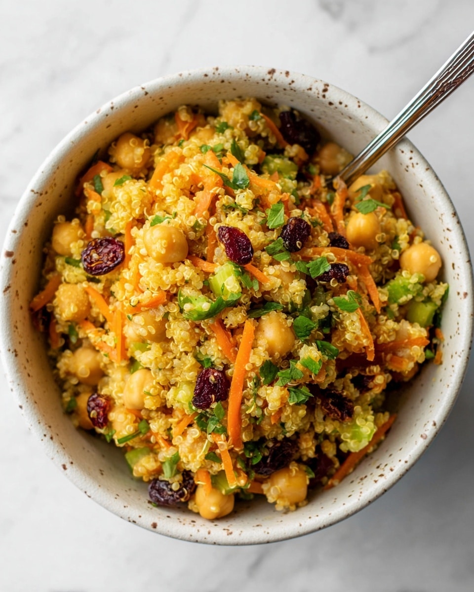 A bowl filled with a colorful quinoa salad, showing a mix of light yellow quinoa grains, round chickpeas, thin orange carrot shreds, small pieces of green celery, and dark red dried cranberries, all topped with bright green fresh herbs. The bowl is white with small brown specks, and a silver spoon rests inside it at the top right. The background is a white marbled texture. photo taken with an iphone --ar 4:5 --v 7