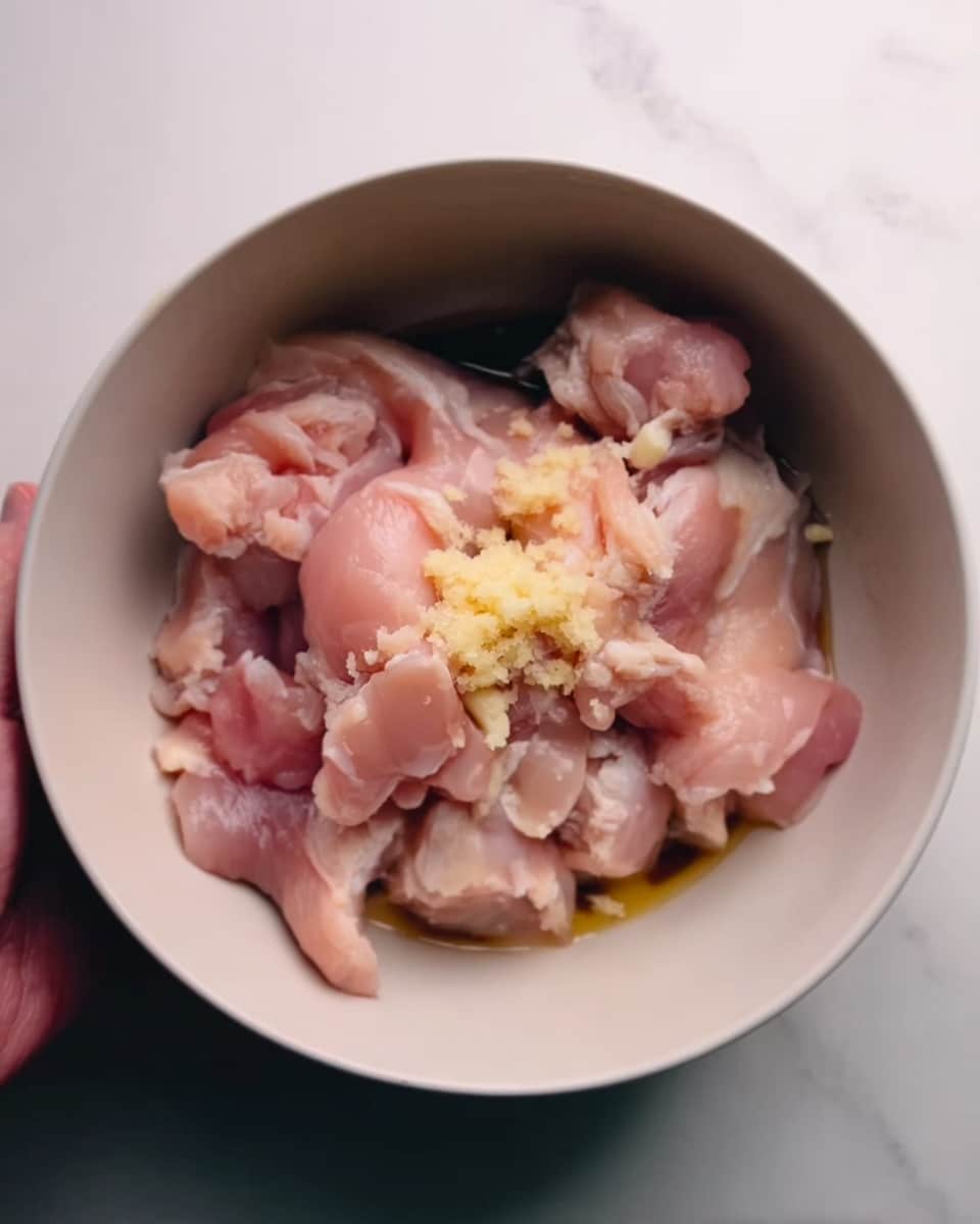 Inside a white bowl, there are several raw pale pink chicken pieces mixed with small bits of light yellow minced garlic on top. The chicken looks fresh and moist with some slightly darker pink areas, and a woman's hand is seen holding the edge of the bowl. Everything is placed on a white marbled surface. Photo taken with an iphone --ar 4:5 --v 7