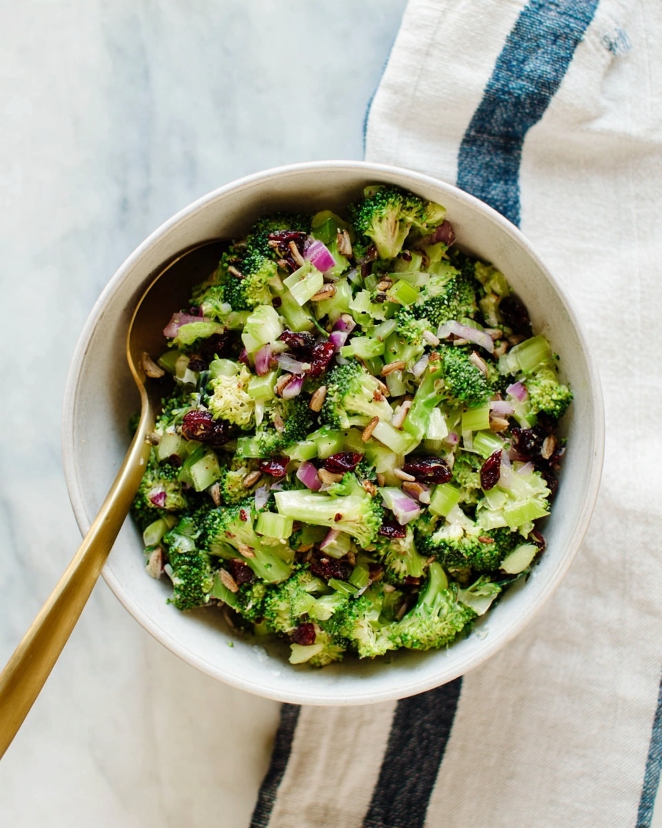 A white bowl filled with a salad made of small green broccoli florets, chopped light green celery, bits of purple-red onion, and small pieces of dark dried cranberries, all mixed with light brown sunflower seeds scattered throughout. There is a gold spoon inside the bowl, resting on the left side. The bowl is placed on a white cloth with a dark blue stripe, lying on a white marbled surface. photo taken with an iphone --ar 4:5 --v 7