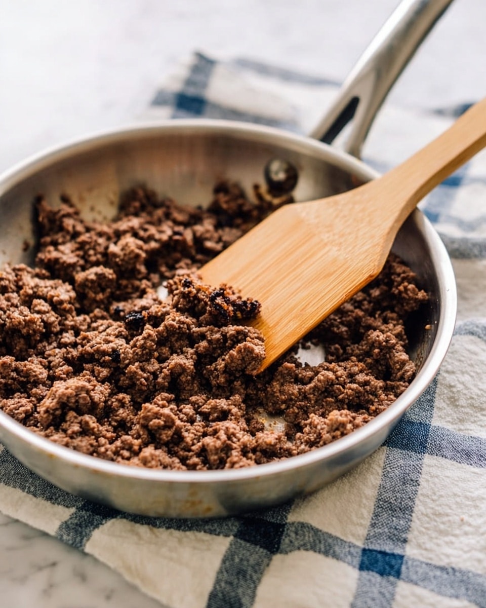 The image shows a stainless steel frying pan filled with cooked ground beef that is dark brown with a crumbly texture. A wooden spatula with a light brown handle rests in the pan, slightly pressing into the beef. The pan is placed on a white and blue checkered cloth on a white marbled surface. The background is softly blurred, focusing on the pan and beef. Photo taken with an iphone --ar 4:5 --v 7